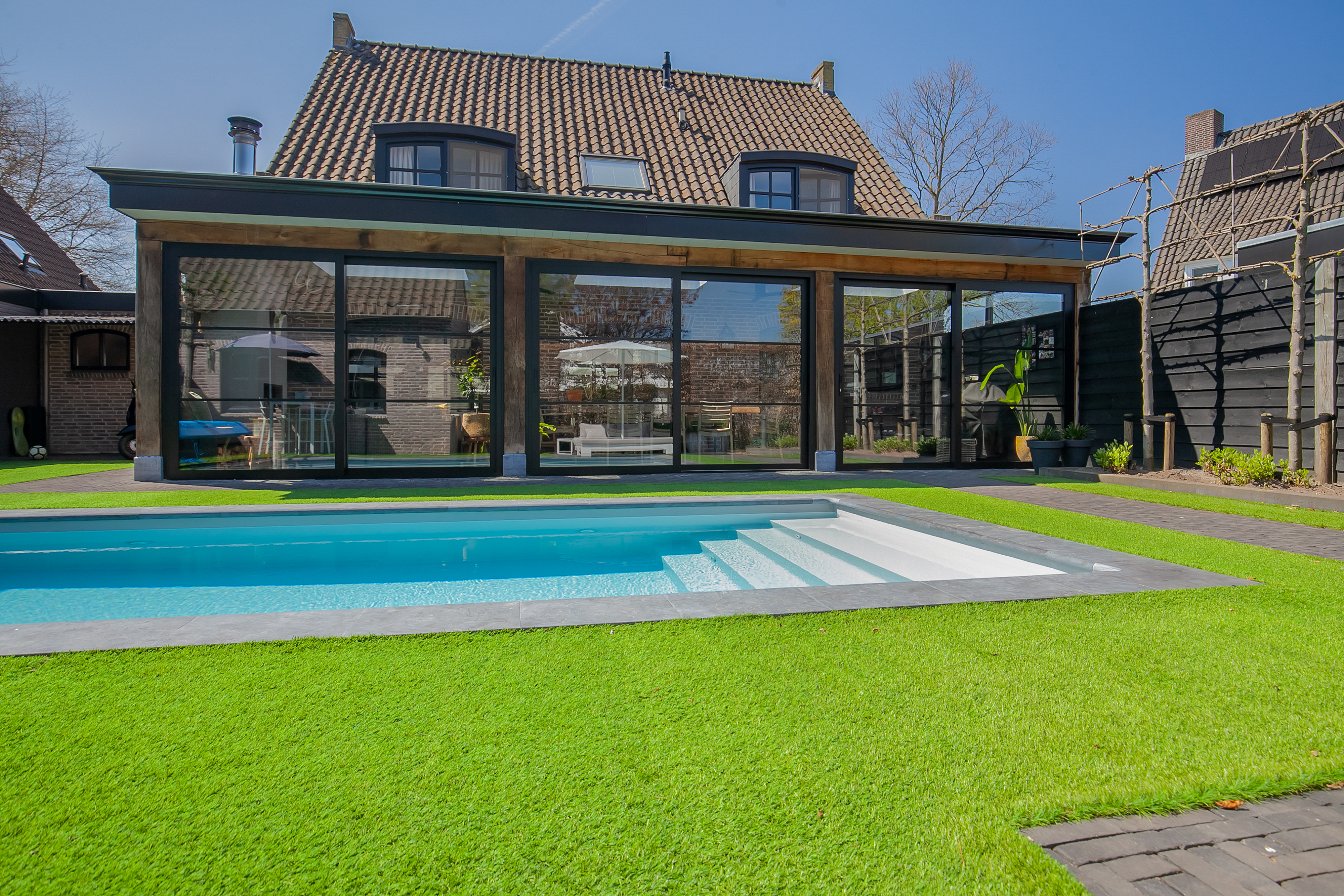 De afbeelding toont een modern huis met grote schuifpuien en deuren die uitkomen op een achtertuin met een zwembad. De kozijnen zijn van donker materiaal, wat een strak contrast geeft met het heldere groene gazon. De bovenverdieping heeft traditionele dakkapellen, wat een klassiek tintje geeft aan het eigentijdse ontwerp.