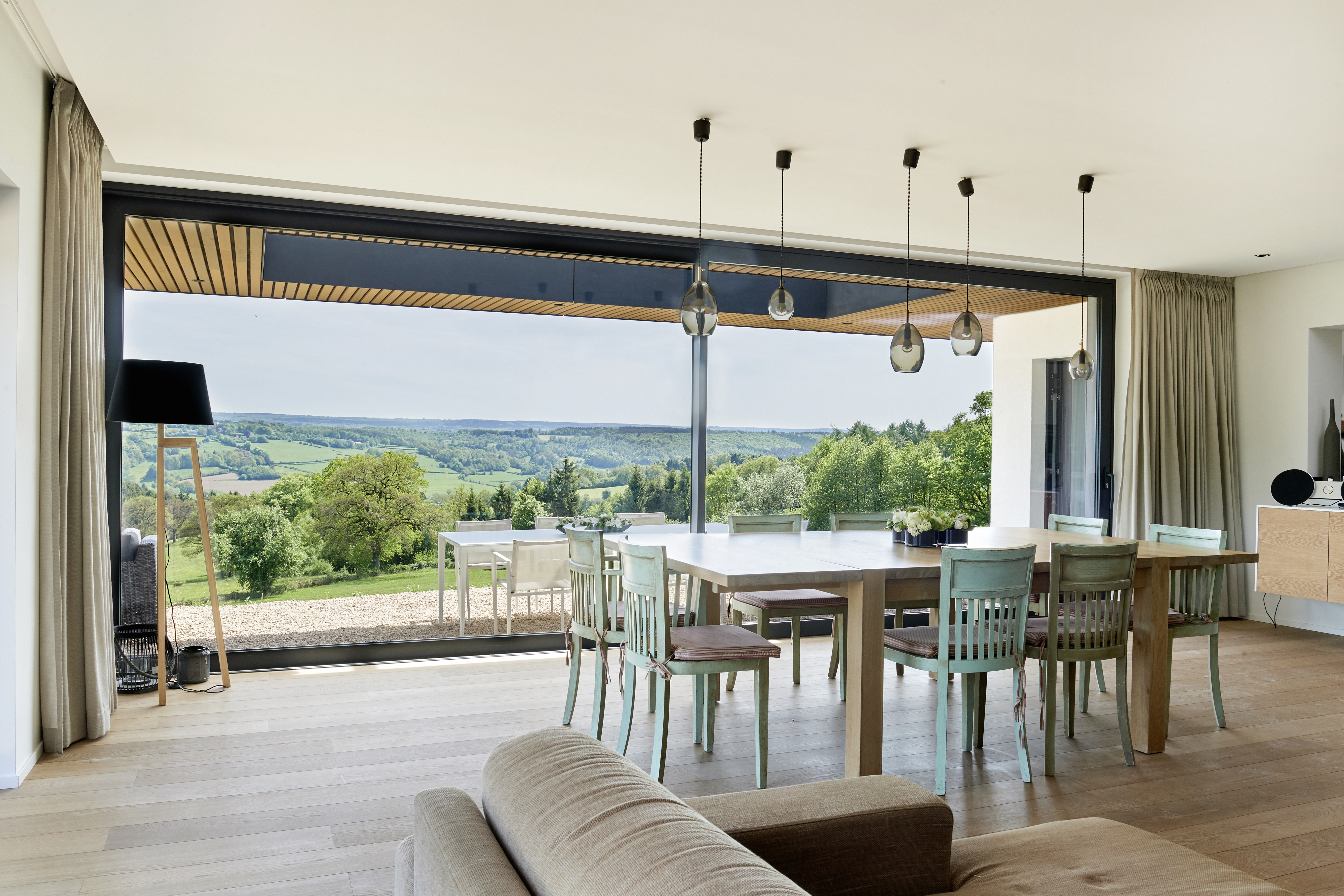 L'image montre un espace de vie moderne avec de grands châssis coulissants qui s'ouvrent sur une vue imprenable sur la campagne. Les châssis laissent entrer une abondance de lumière naturelle, créant une atmosphère lumineuse et aérée. Le design épuré des portes coulissantes complète l'intérieur contemporain, renforçant la connexion entre les espaces intérieurs et extérieurs.