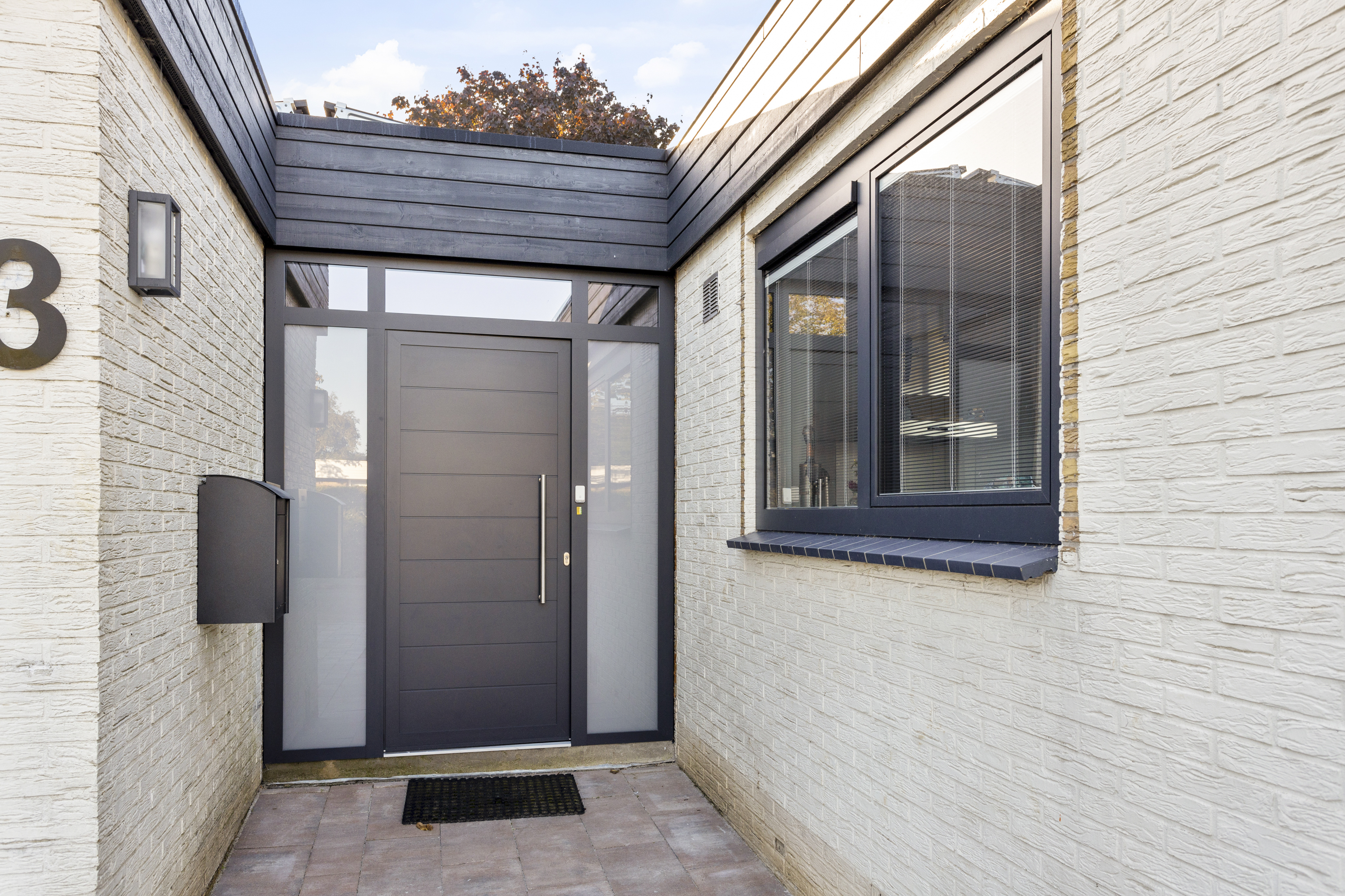 De afbeelding toont een moderne entree met een strakke zwarte deur en grote kozijnen. De deur heeft horizontale panelen en een lange verticale handgreep, geflankeerd door matglazen panelen. De aangrenzende kozijnen zijn groot, met zwarte lijsten, waardoor er veel licht naar binnen valt. De buitenmuren zijn van lichtgekleurde baksteen, wat contrasteert met de donkere lijsten.