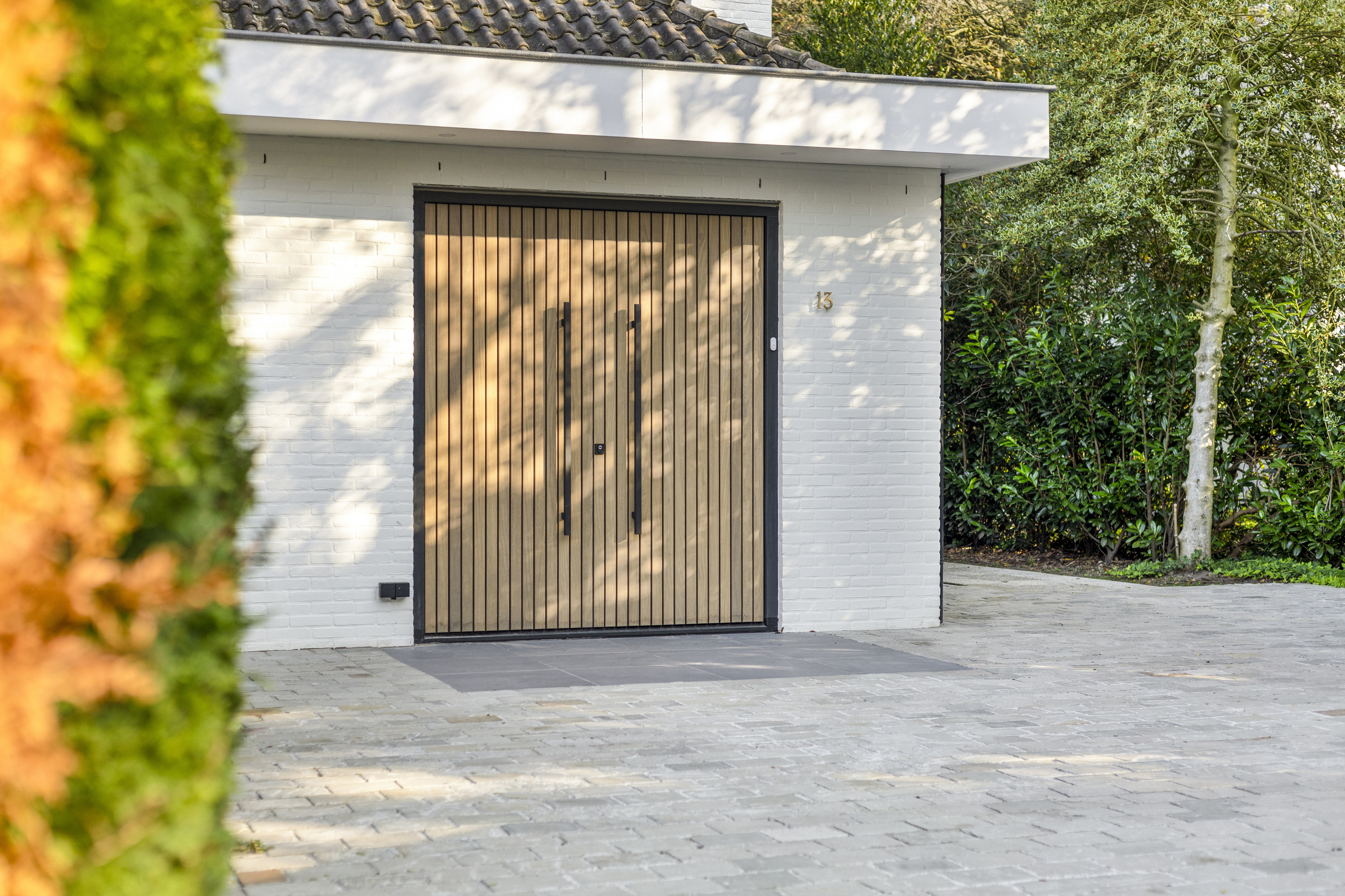 De afbeelding toont een moderne houten garagedeur in een witte bakstenen muur. De deur heeft verticale houten latten en strakke zwarte handgrepen, wat een eigentijdse uitstraling geeft. De omgeving is bestraat met lichtgekleurde stenen en er is groen zichtbaar op de achtergrond.