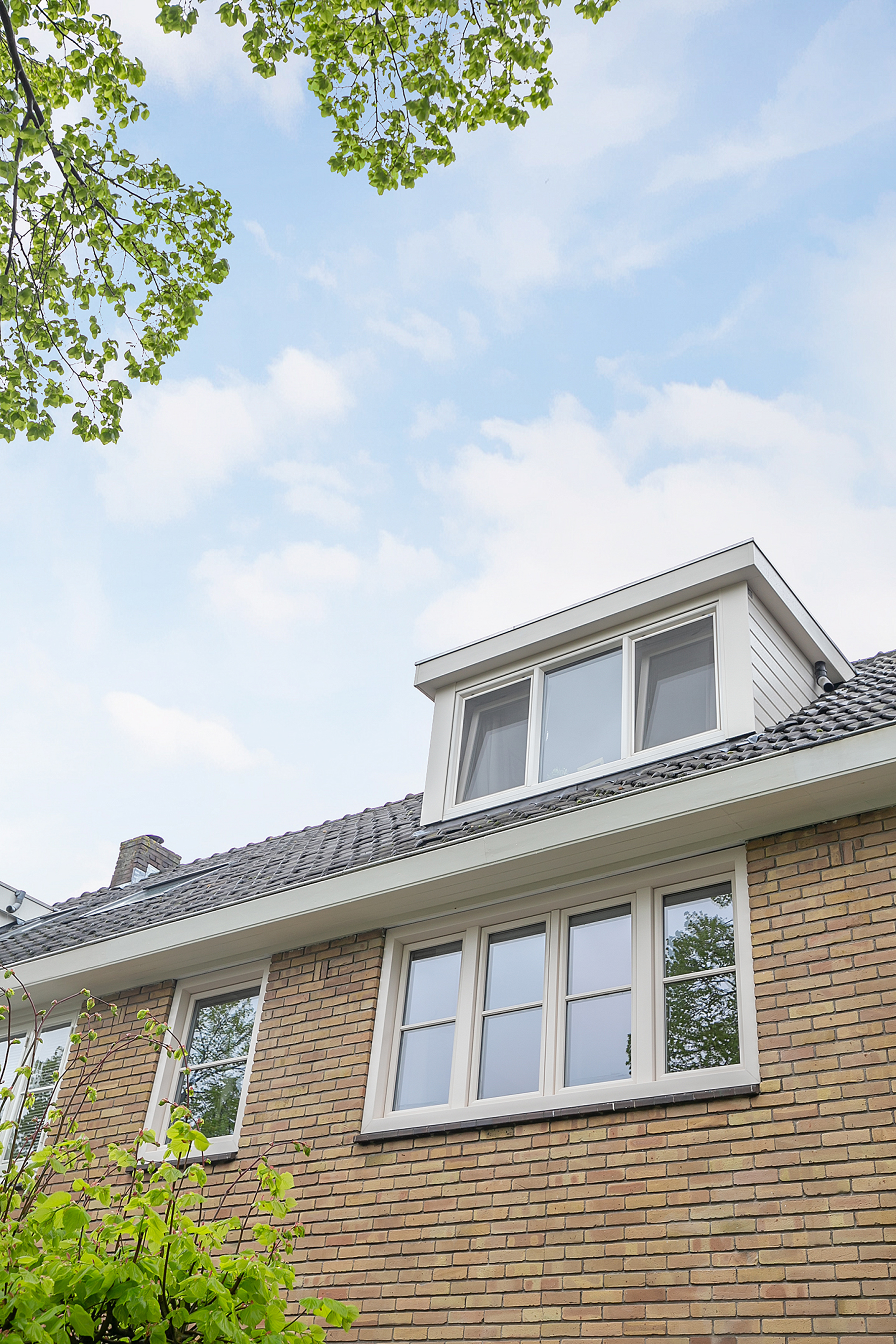 De afbeelding toont een bakstenen huis met een zadeldak. De kozijnen zijn wit, met een set van drie op de dakkapel en twee sets dubbele kozijnen eronder. Het ontwerp is klassiek en symmetrisch, wat de esthetiek van het huis versterkt.