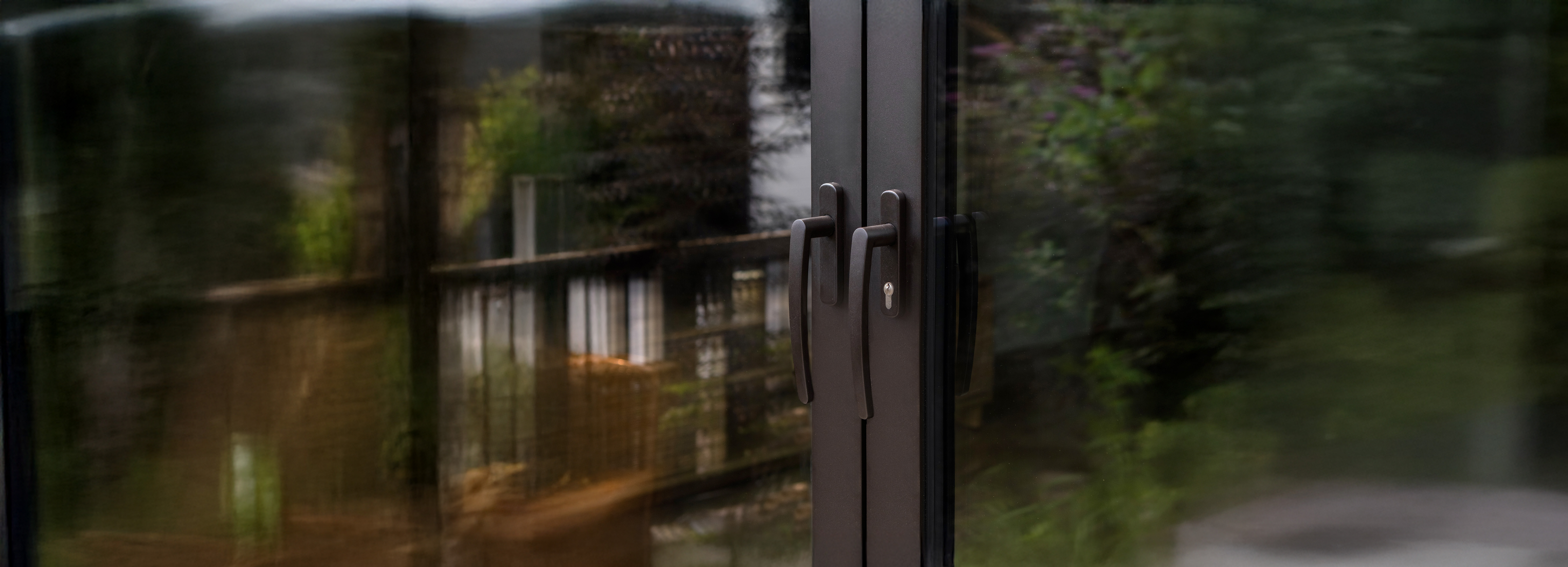 De afbeelding toont een moderne schuifpui met strakke zwarte handgrepen. Het glas weerspiegelt een wazig beeld van een tuin, wat een naadloze verbinding tussen binnen en buiten creëert. Het ontwerp van de pui is minimalistisch, met nadruk op functionaliteit en stijl.