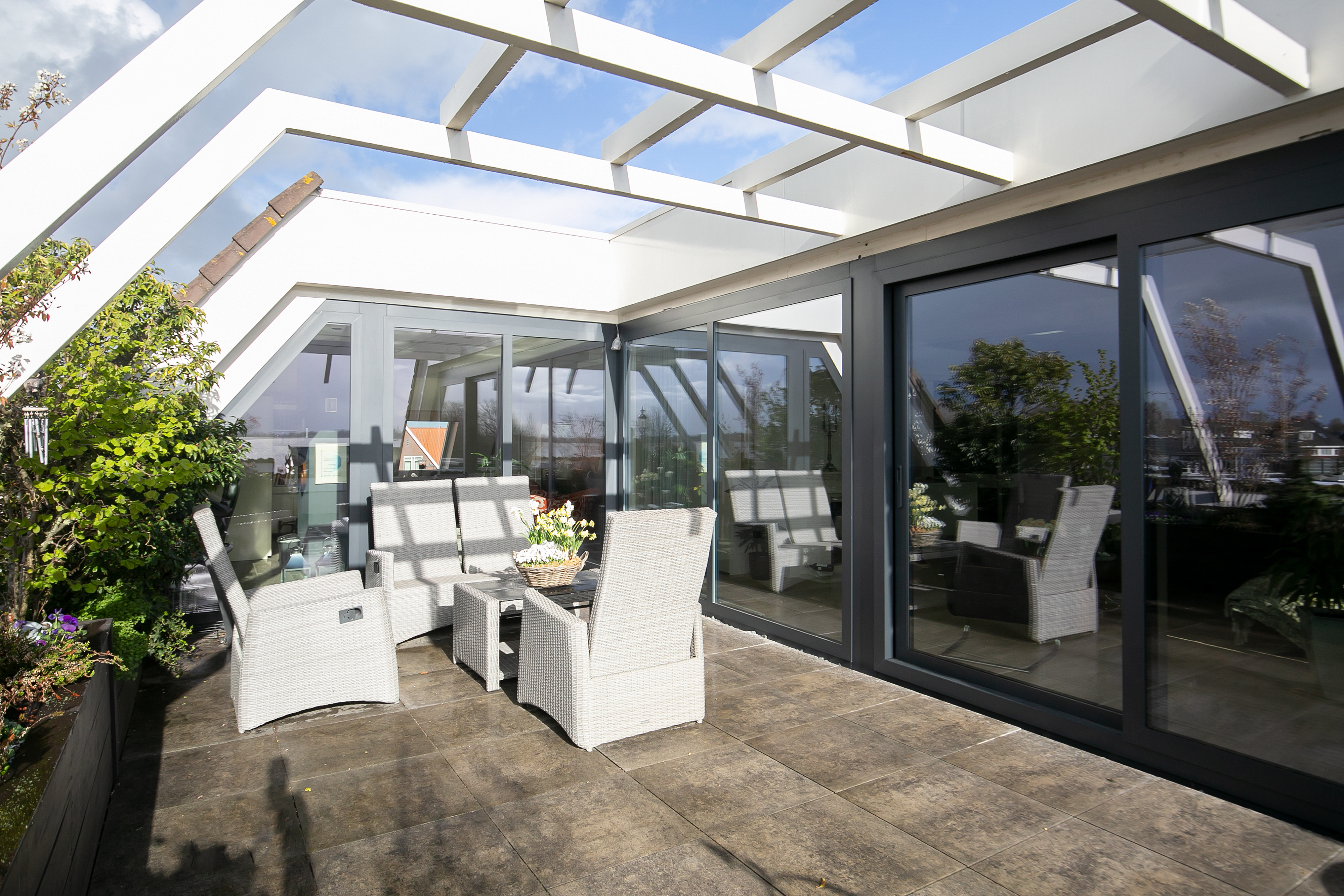 De afbeelding toont een moderne buitenpatio met grote glazen schuifpuien en kozijnen. De kozijnen en schuifpuien hebben een strak, donker metalen frame, wat een eigentijdse uitstraling geeft. De schuifpuien openen naar een ruim terras, wat zorgt voor een naadloze overgang tussen binnen en buiten. Het glas weerspiegelt de omliggende groen en lucht, wat bijdraagt aan de open en luchtige sfeer.