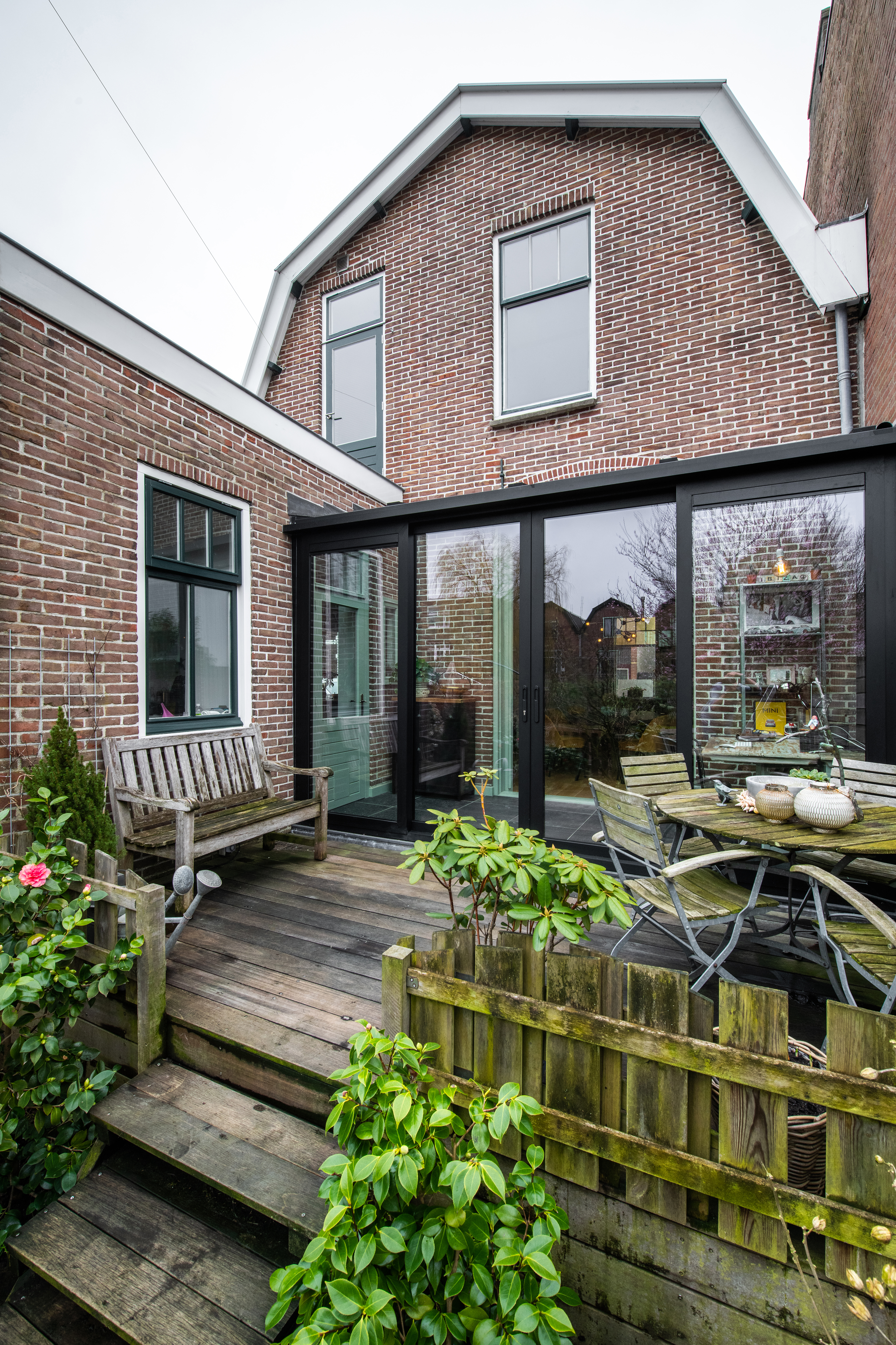 De afbeelding toont een charmant bakstenen huis met grote kozijnen en een schuifpui die leidt naar een houten terras. De kozijnen zijn modern en laten veel natuurlijk licht binnen. De schuifpui heeft een zwart frame, wat een eigentijdse uitstraling geeft aan de traditionele bakstenen gevel.