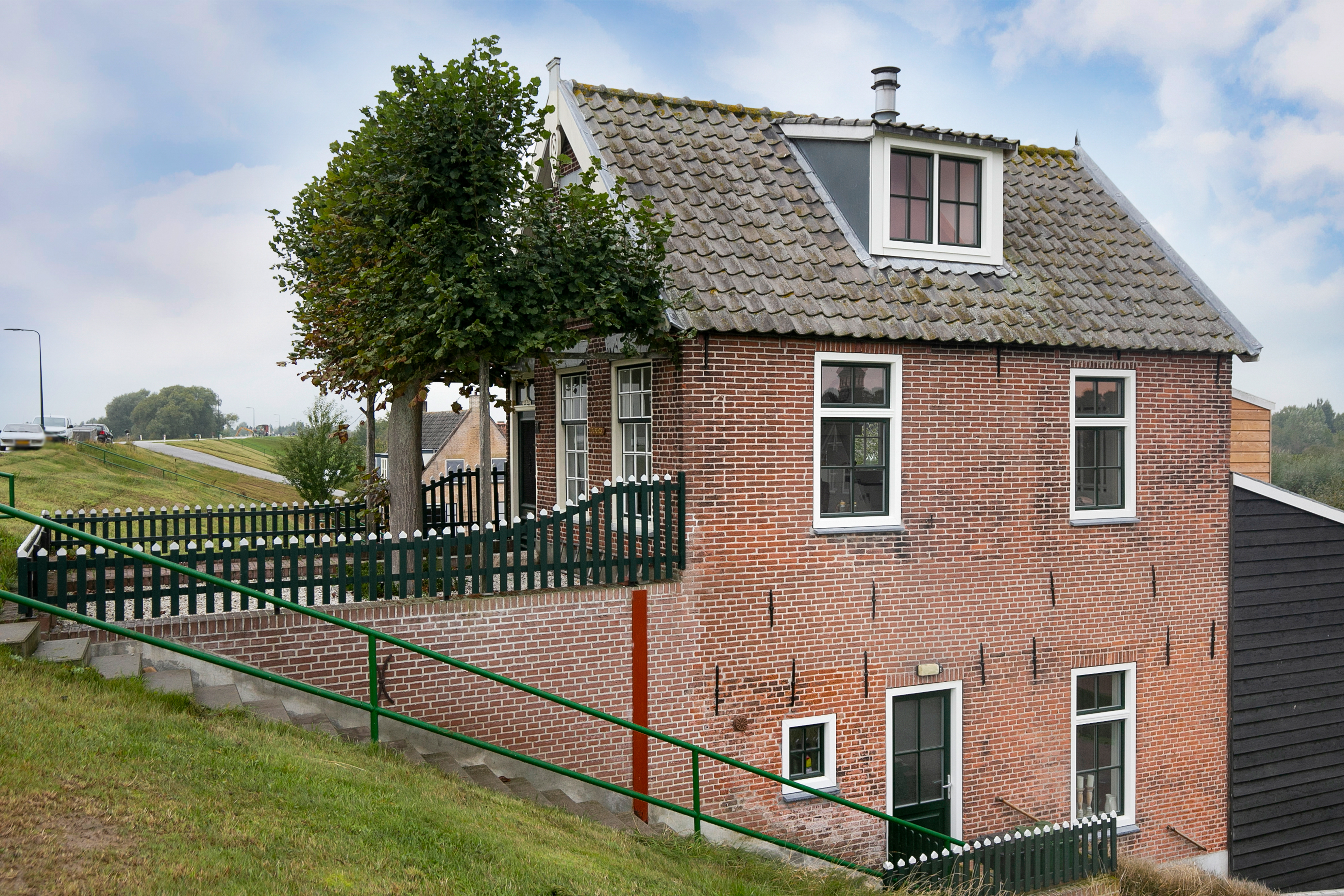 Het huis heeft traditionele baksteen met meerdere kozijnen en een deur. De kozijnen zijn symmetrisch geplaatst, met witte lijsten die afsteken tegen de rode baksteen. De voordeur is donker en past bij het geheel.