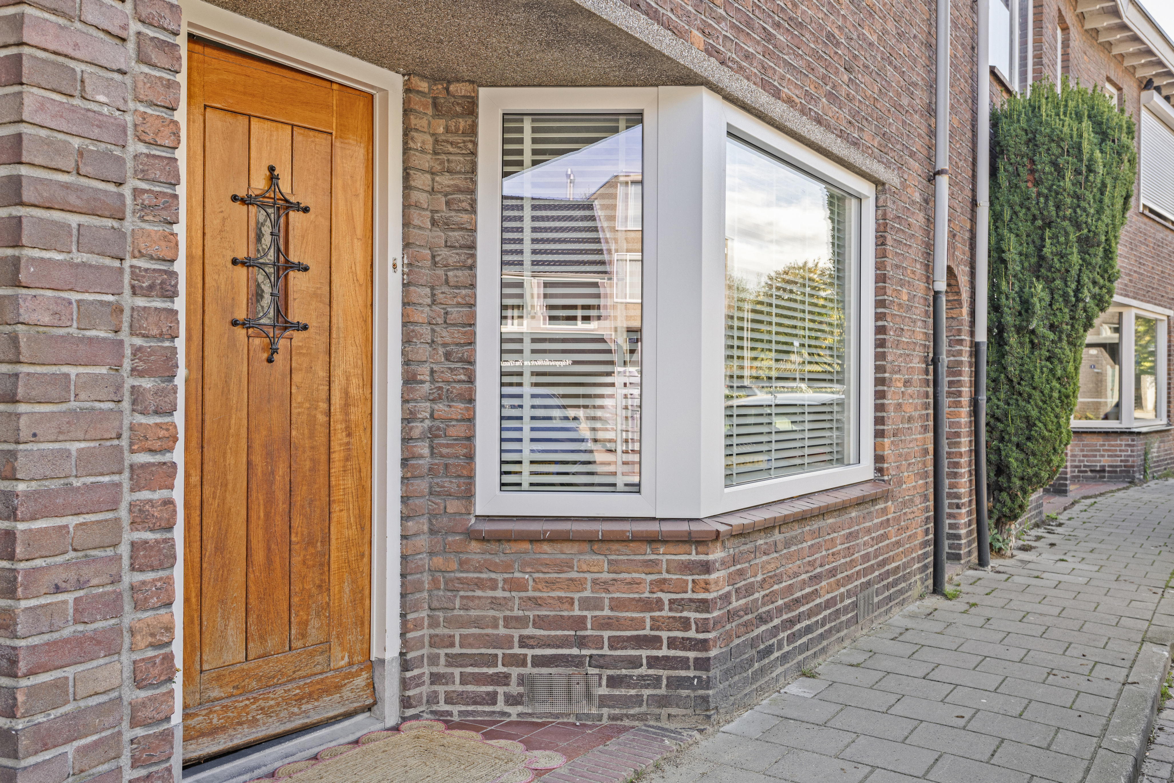 De afbeelding toont een traditioneel bakstenen huis met een houten voordeur met decoratief smeedwerk. Naast de deur zijn er twee grote kozijnen met witte kozijnen en horizontale jaloezieën die het straatbeeld weerspiegelen.