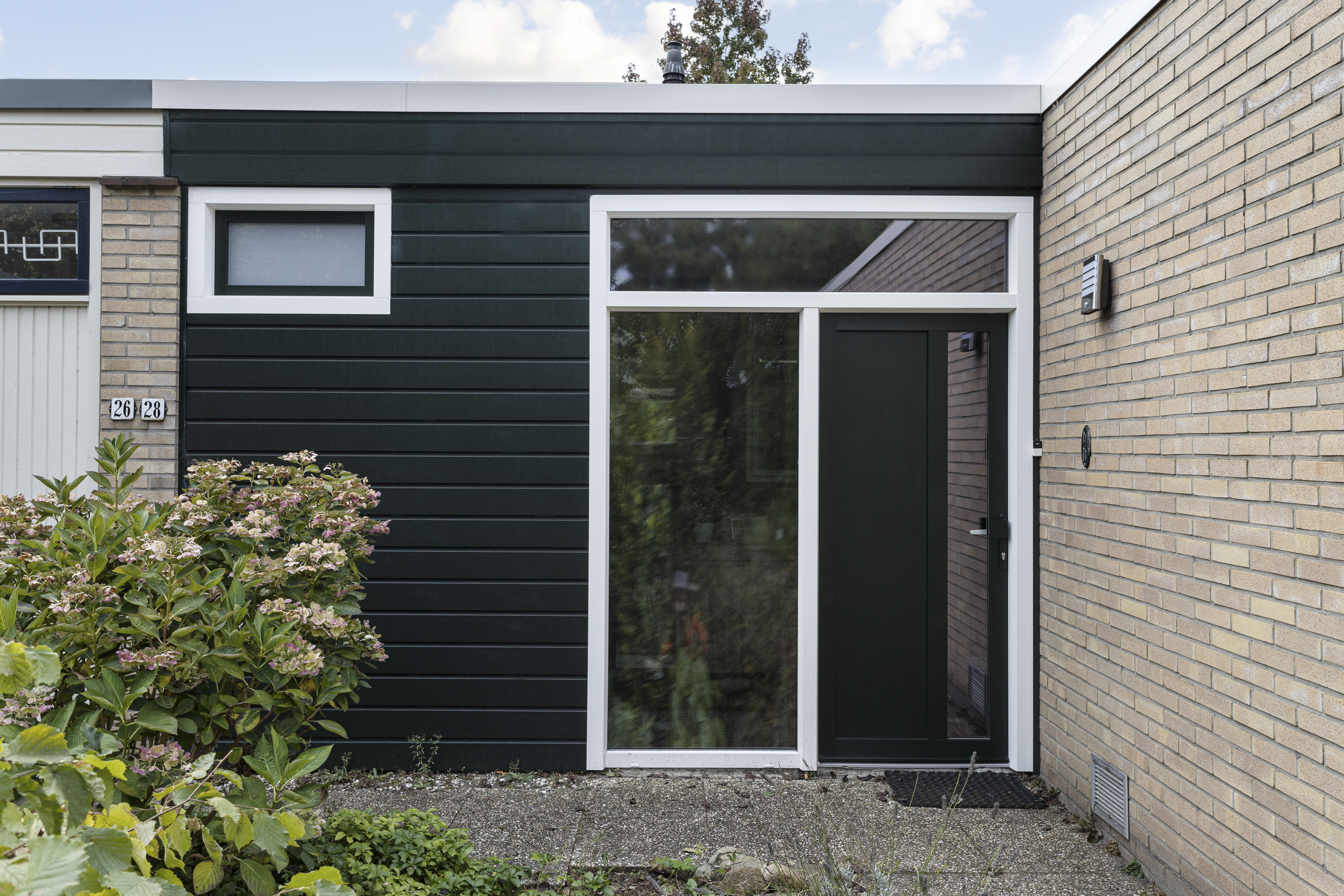 De afbeelding toont een moderne gevel met een donkergroene deur en een groot kozijn ernaast. Het kozijn is rechthoekig en biedt een blik naar binnen. Boven de deur is er een kleiner kozijn, wat bijdraagt aan het eigentijdse ontwerp. De omliggende muren zijn van lichtgekleurde bakstenen, wat contrasteert met de donkere elementen.