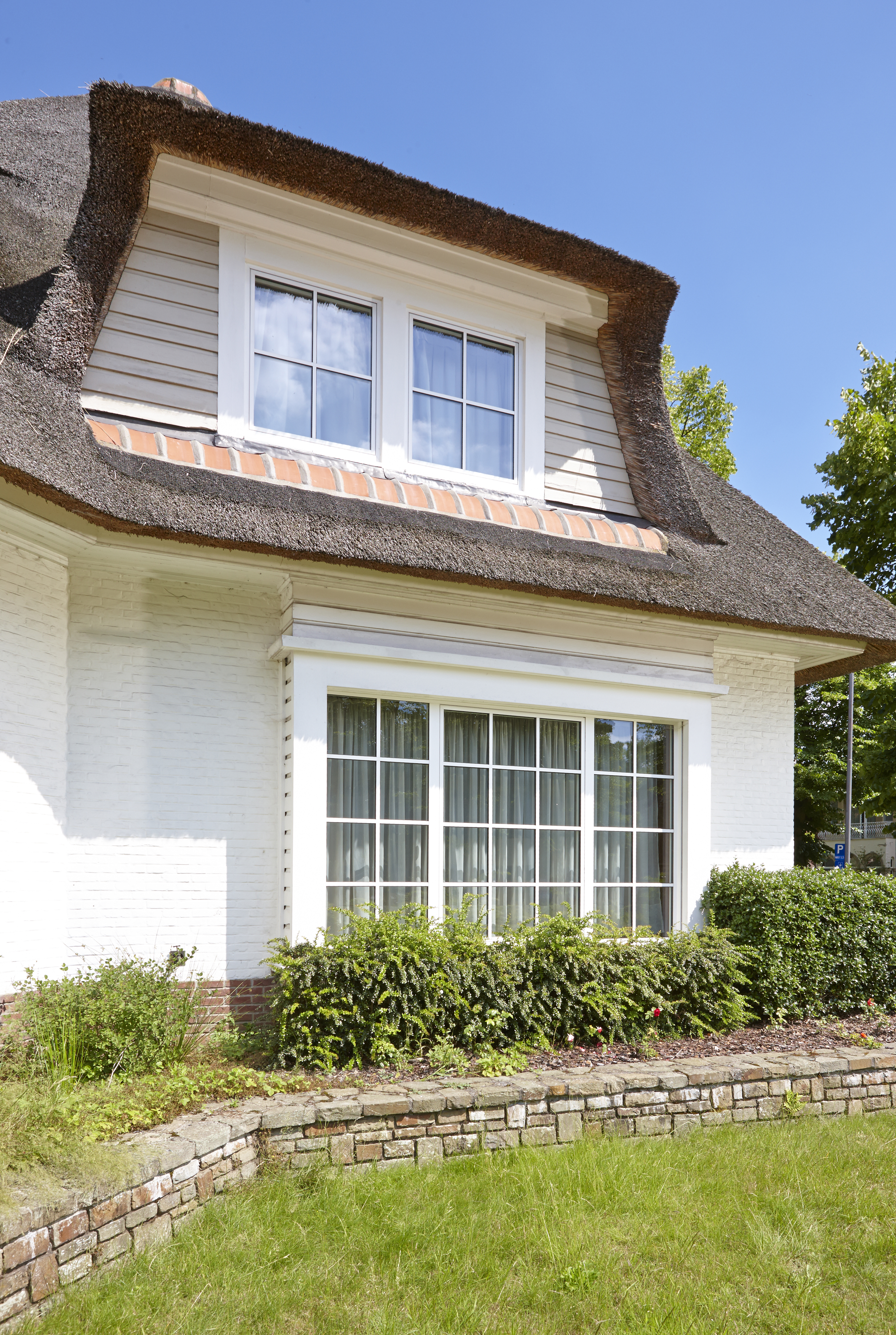 L'image montre une maison charmante avec un toit de chaume. La façade présente de grandes châssis à cadre blanc qui laissent entrer beaucoup de lumière naturelle. À l'étage supérieur, il y a deux plus petits châssis, également avec des cadres blancs, intégrés dans une lucarne. Les châssis sont disposés symétriquement, renforçant l'attrait esthétique de la maison.