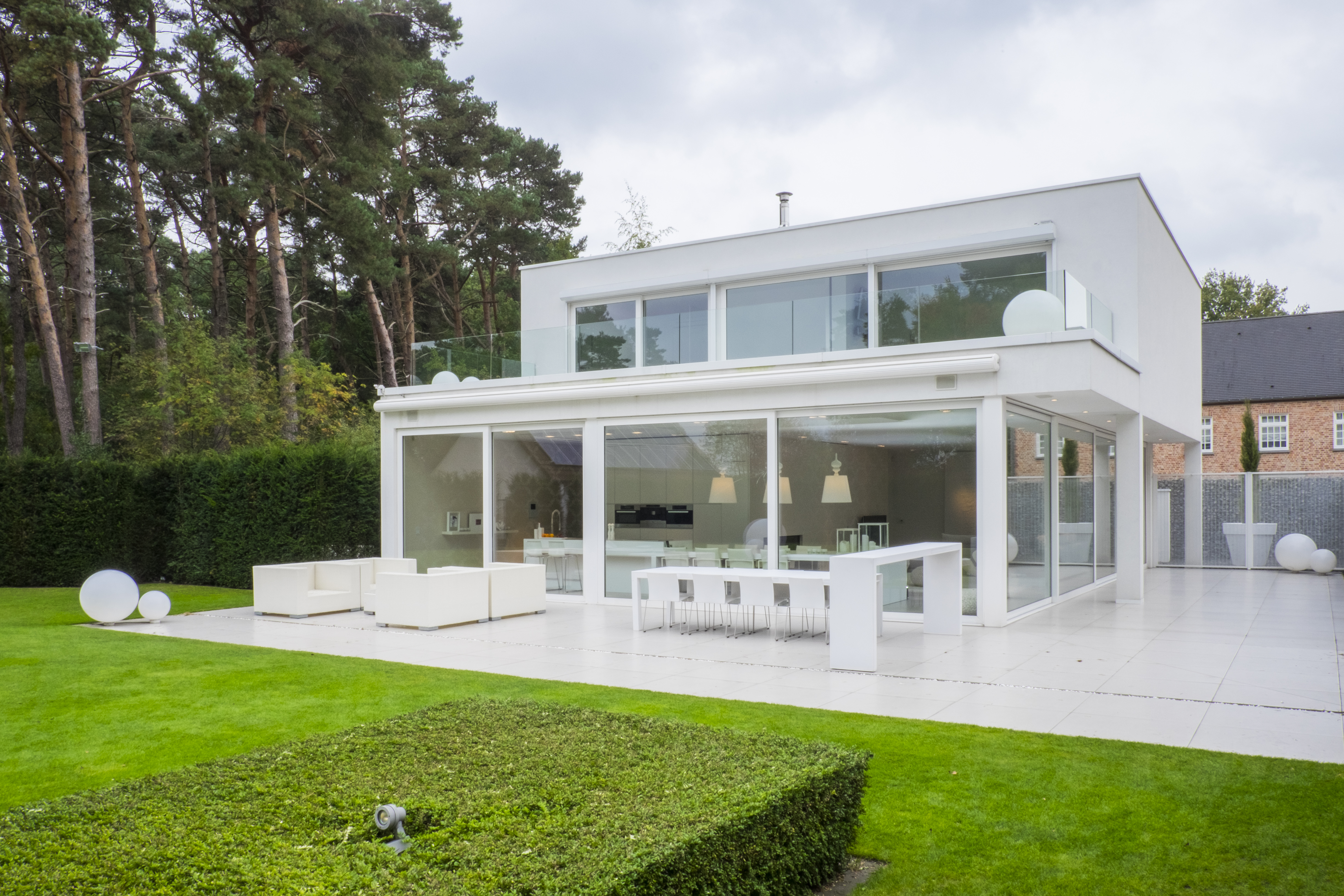 Het moderne huis heeft grote schuiframen en deuren, waardoor er een naadloze verbinding ontstaat tussen binnen en buiten. De benedenverdieping is omgeven door uitgestrekte glazen panelen, die natuurlijk licht binnenlaten. De bovenverdieping heeft ook brede ramen, wat het eigentijdse ontwerp versterkt en panoramische uitzichten biedt op het omliggende groen.