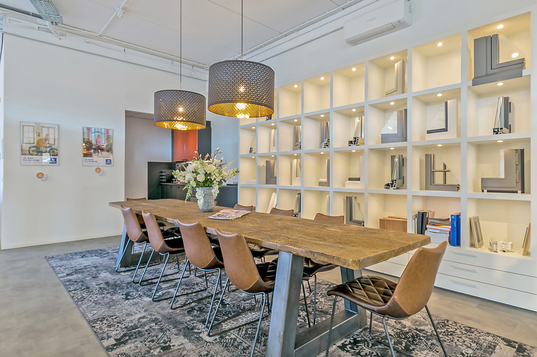 De afbeelding toont een moderne showroom met een focus op kozijnen en deuren. Een grote houten tafel is omringd door stijlvolle stoelen, en de muur heeft een display van verschillende kozijn- en deurprofielen. De ruimte is goed verlicht, wat het strakke ontwerp en de functionaliteit van de producten benadrukt.