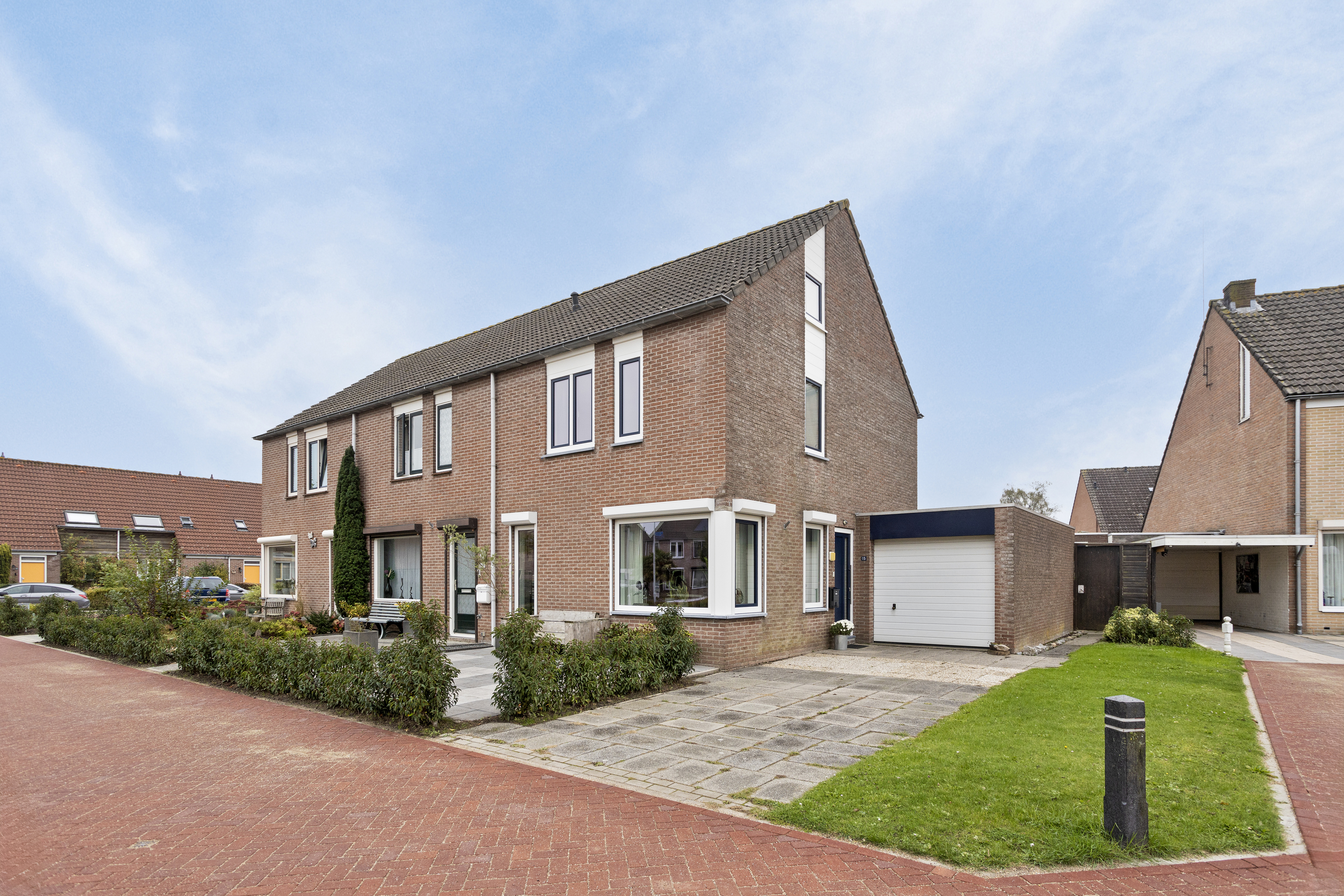 De afbeelding toont een twee-onder-een-kapwoning met een bakstenen gevel. Het huis heeft meerdere grote kozijnen, waaronder erkers op de begane grond, die veel natuurlijk licht binnenlaten. De voordeur is centraal geplaatst, geflankeerd door kozijnen. De garagedeur is wit en grenst aan de hoofdingang.