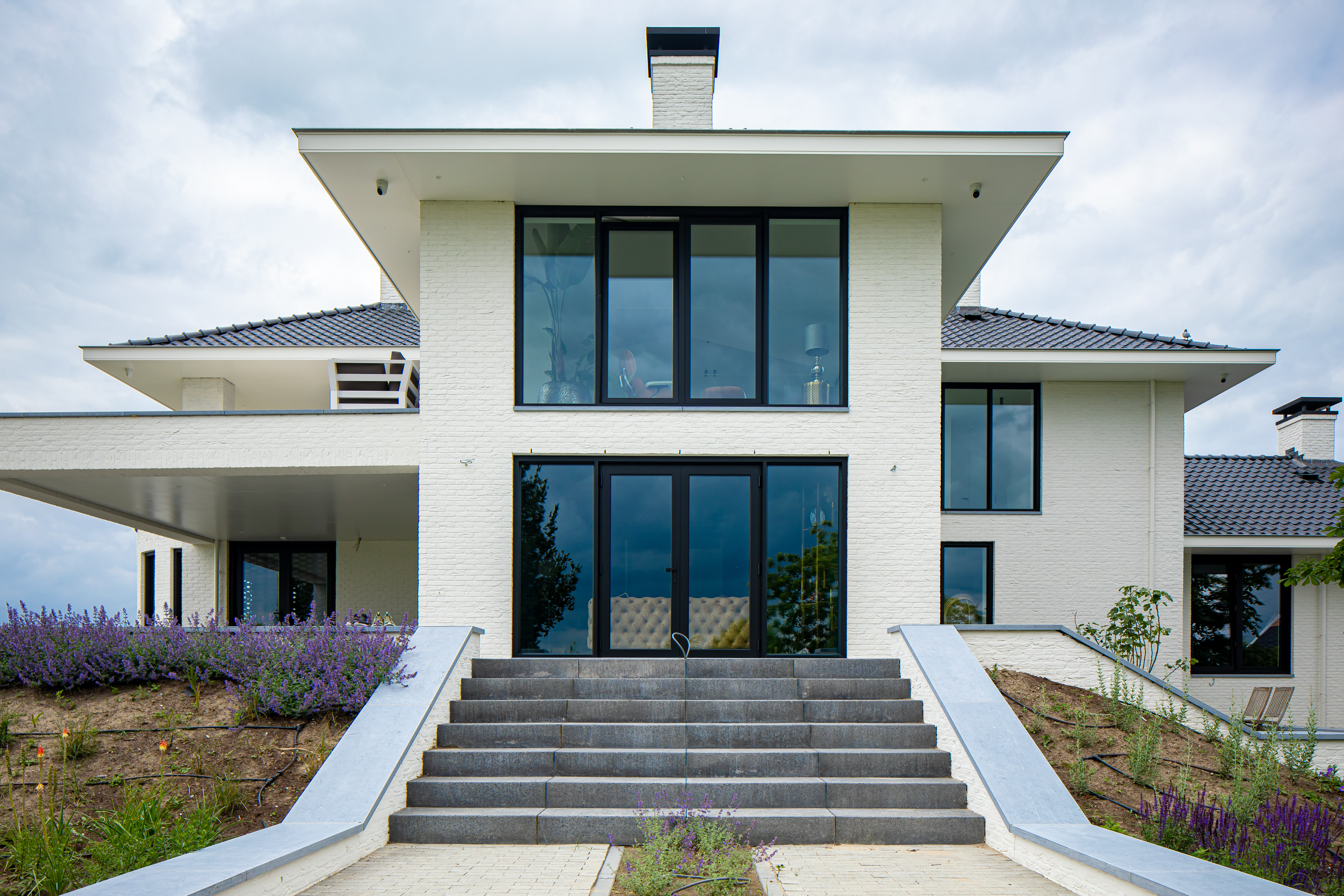 Het moderne huis heeft grote, zwartomrande kozijnen en een centrale glazen deur, wat een strakke en eigentijdse uitstraling geeft. De kozijnen zijn symmetrisch geplaatst, wat het architectonische ontwerp versterkt. De entree wordt geaccentueerd door brede treden die naar de deur leiden, wat een uitnodigende gevel creëert.