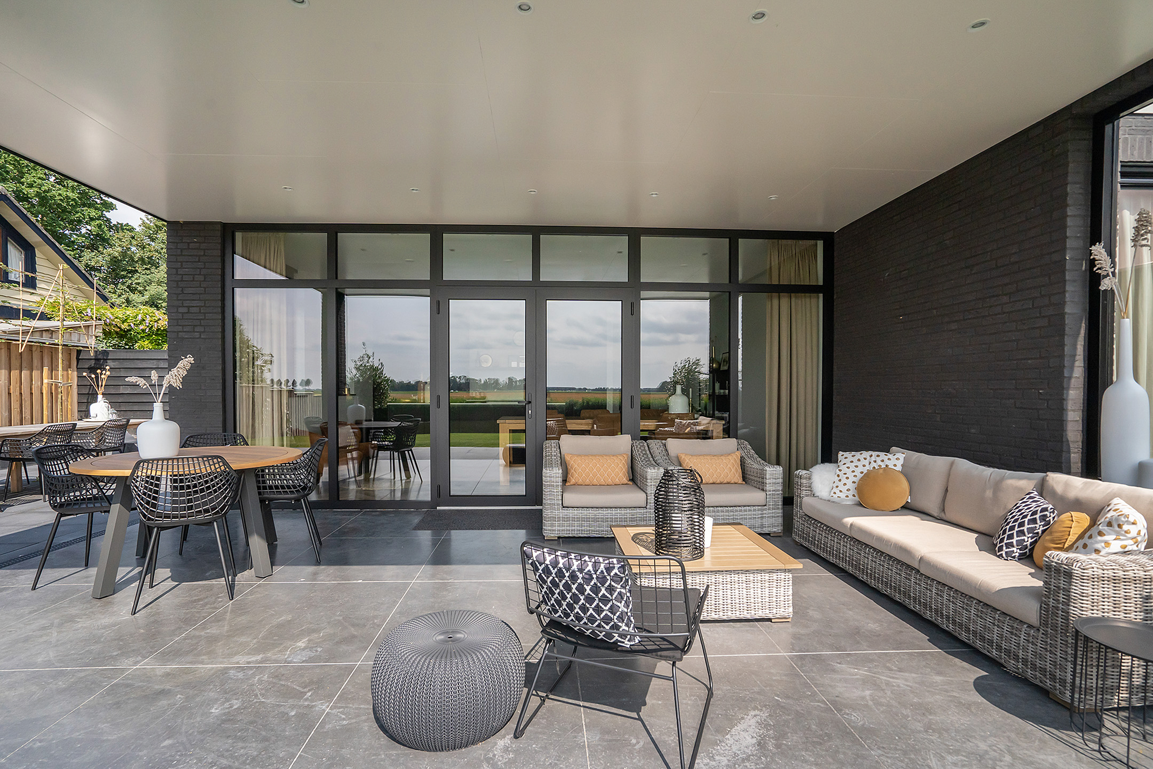 De afbeelding toont een moderne patio met grote schuifpuien en deuren. De glazen panelen zorgen voor een naadloze overgang tussen binnen en buiten, waardoor natuurlijk licht de ruimte binnenstroomt. De kozijnen zijn strak en donker, wat goed past bij het eigentijdse ontwerp van de patio.