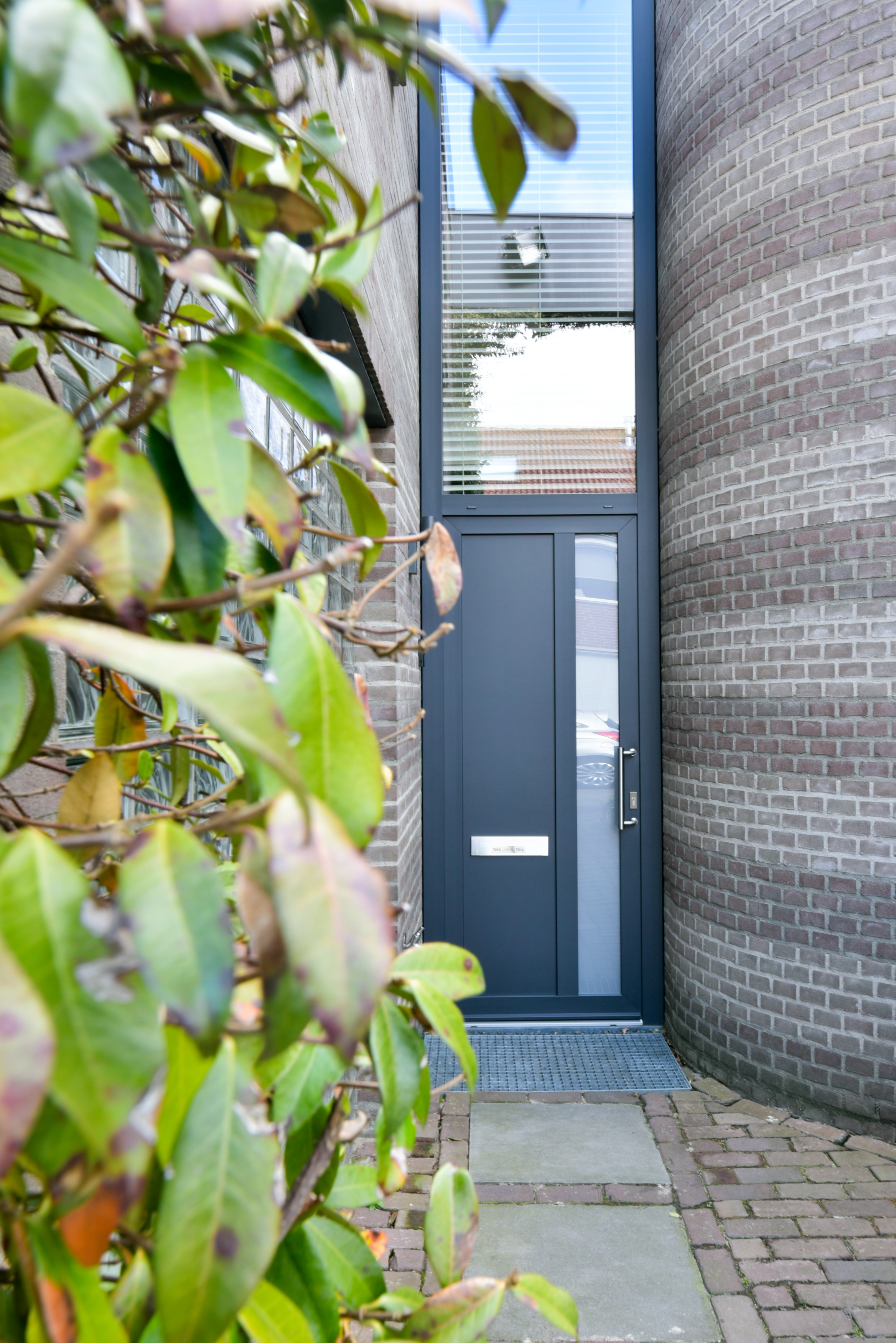 De afbeelding toont een moderne entree met een strak, donkergekleurde deur en een hoog kozijn erboven. De deur heeft een metalen handgreep en een brievenbus, wat het eigentijdse ontwerp aanvult. Het kozijn heeft horizontale jaloezieën, die privacy bieden terwijl er licht binnenkomt. Het omliggende metselwerk voegt een klassiek tintje toe aan de moderne elementen.