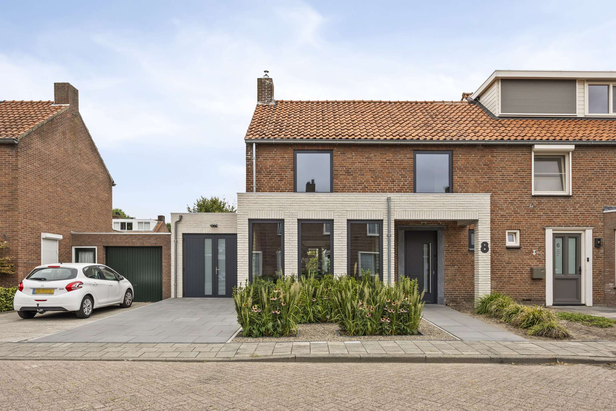 Het huis heeft moderne kozijnen en deuren met strakke, donkere lijsten. De grote kozijnen laten veel natuurlijk licht binnen, wat de bakstenen gevel versterkt. De voordeur is stijlvol en past bij het eigentijdse ontwerp.