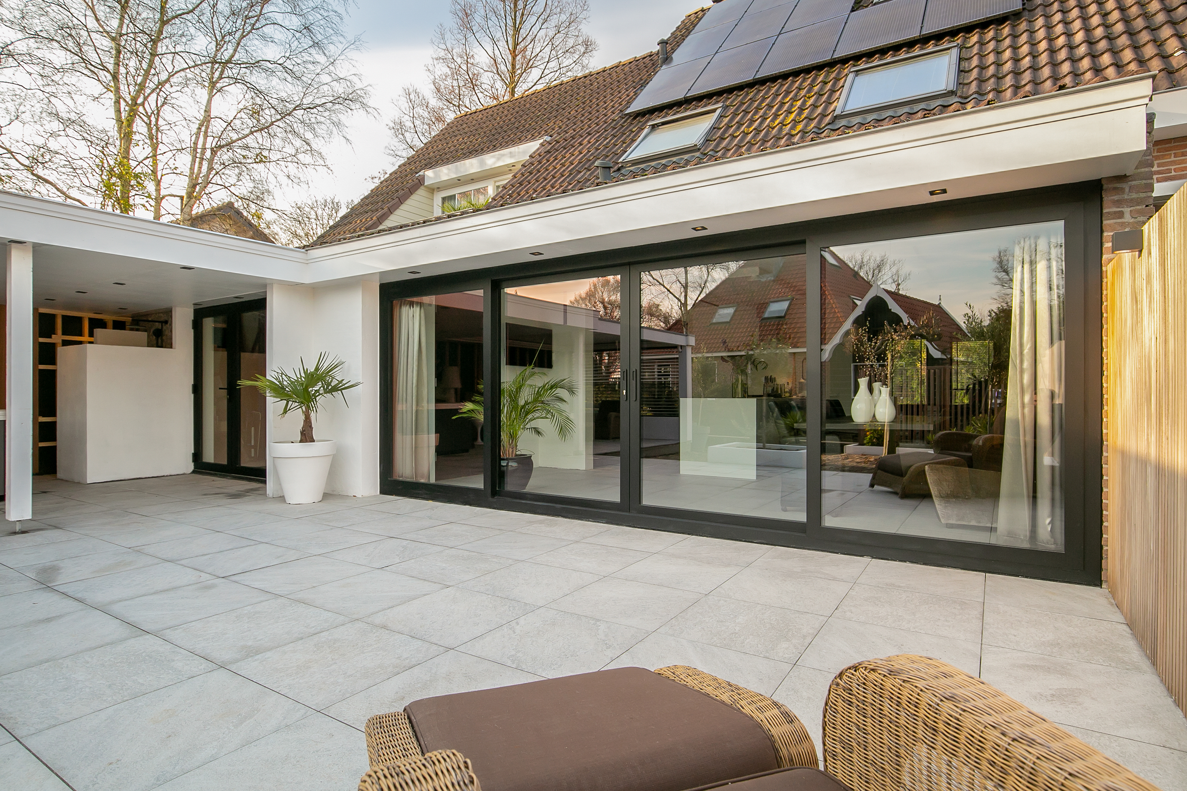 De afbeelding toont een modern huis met grote schuifpuien die leiden naar een ruime patio. De puien hebben een strak zwart frame dat past bij het eigentijdse ontwerp. Bovenop het dak zijn er dakramen die natuurlijk licht binnenlaten. De patio is betegeld met grote tegels, wat de buitenruimte vergroot.