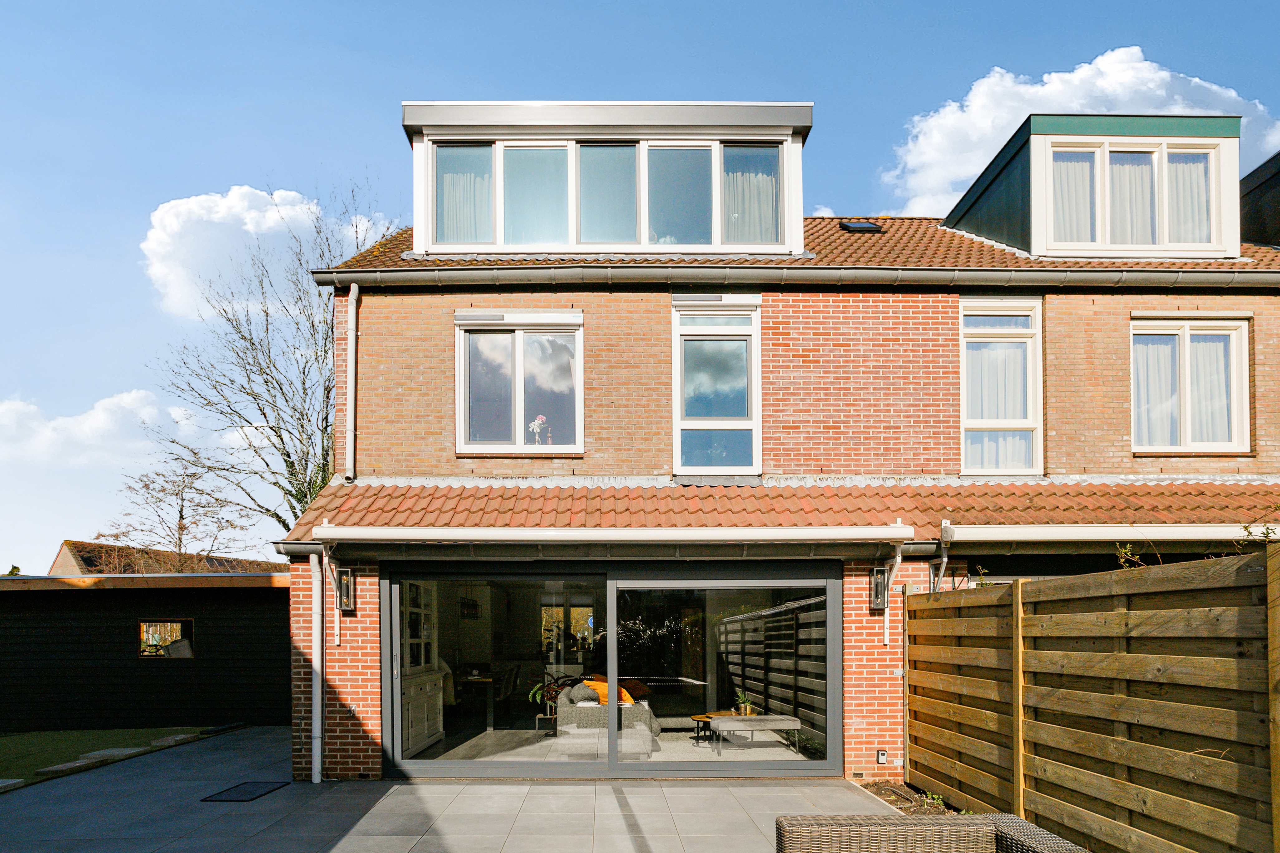 Het huis heeft een modern ontwerp met grote kozijnen en een schuifpui. De bovenverdieping heeft meerdere kozijnen die natuurlijk licht binnenlaten. De begane grond heeft een brede schuifpui die naar het terras leidt, wat binnen-buiten leven bevordert.