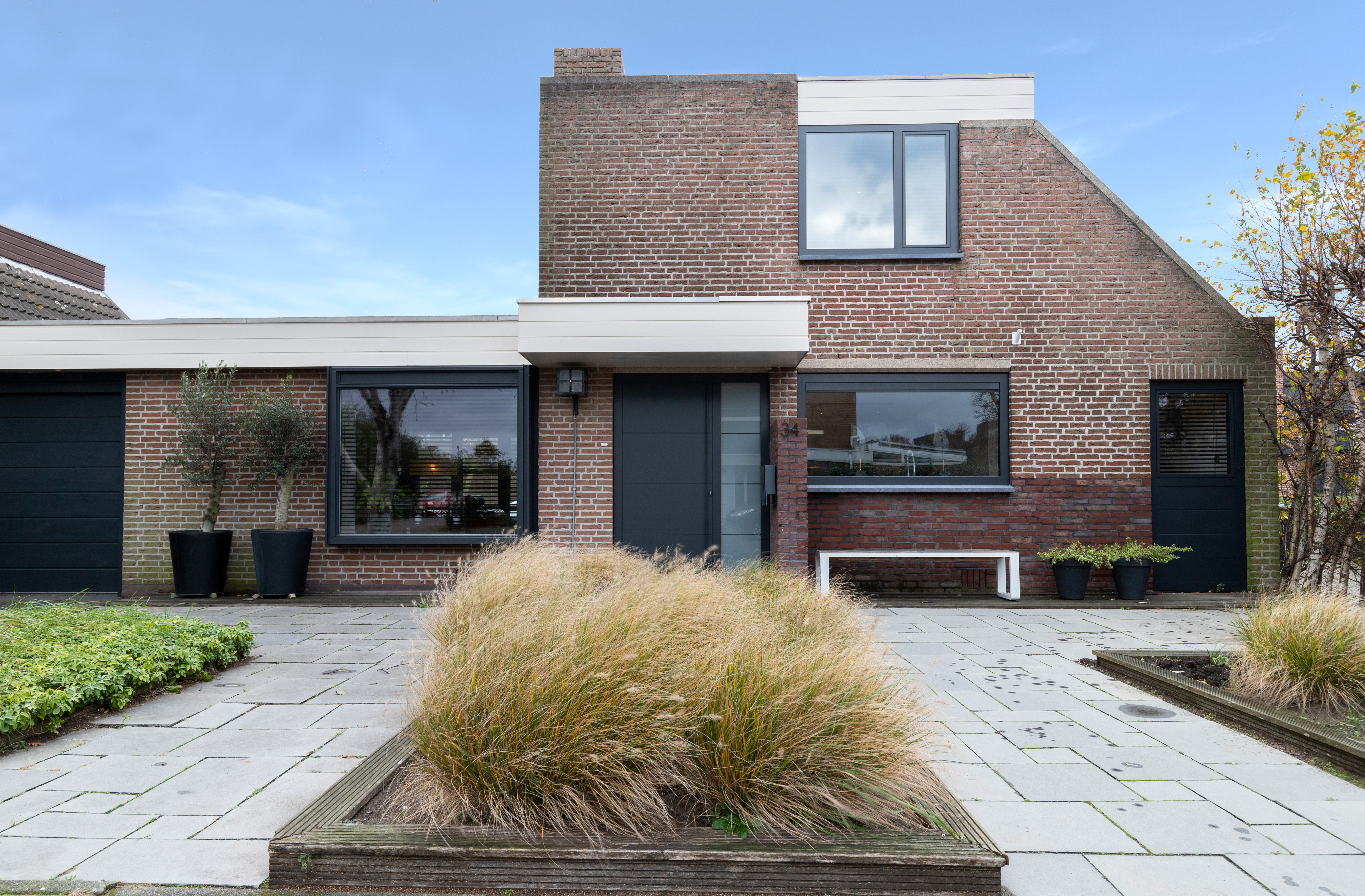 Het huis heeft een modern ontwerp met grote rechthoekige kozijnen en een strakke zwarte deur. De kozijnen zijn omlijst met donker materiaal, wat mooi contrasteert met de bakstenen gevel. De voordeur is centraal geplaatst, geflankeerd door twee kozijnen, wat een evenwichtige uitstraling geeft.