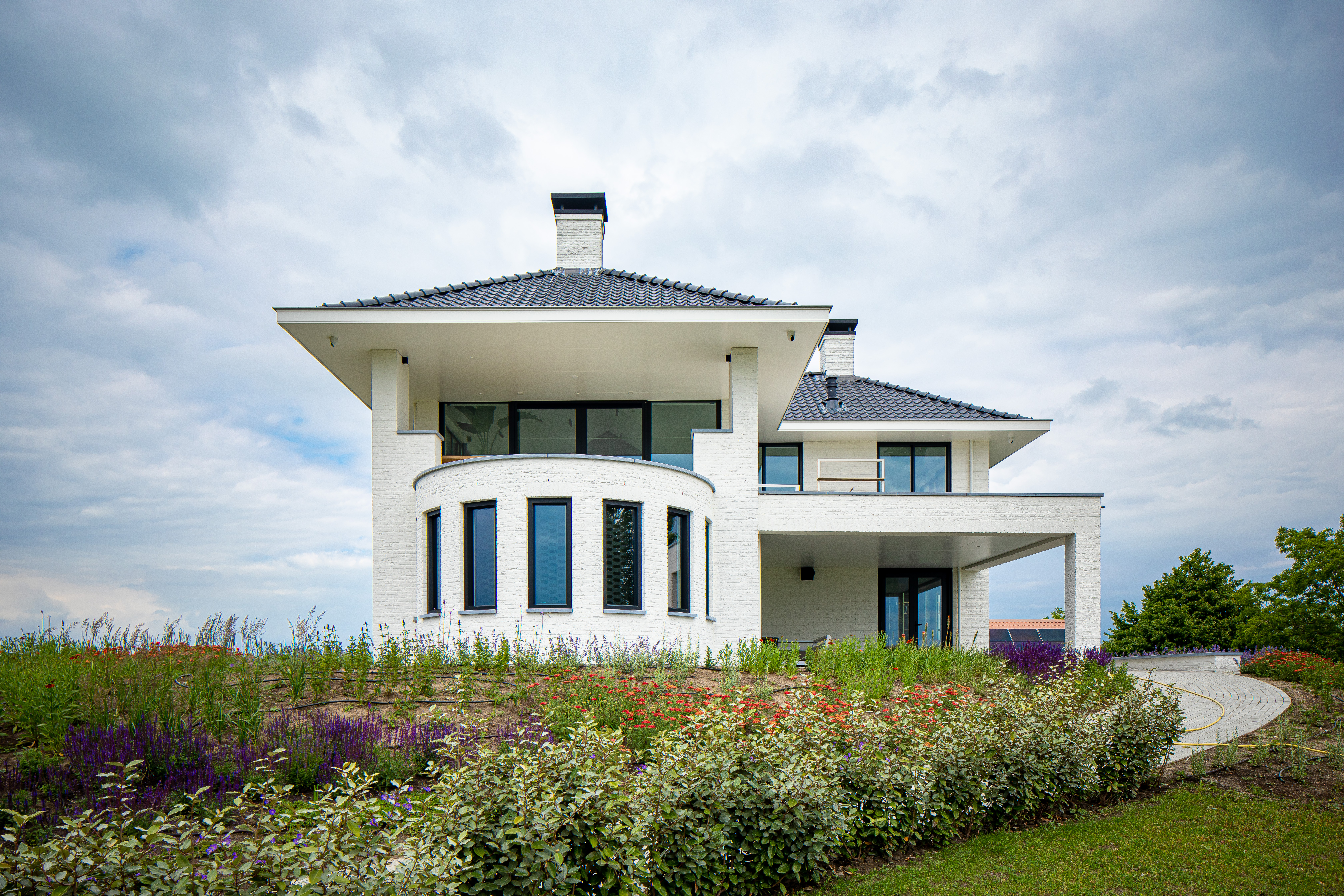 Het moderne huis heeft grote, strakke kozijnen en deuren die het eigentijdse ontwerp versterken. De voorkant toont hoge, smalle kozijnen die veel natuurlijk licht binnenlaten, terwijl de hoofdingang wordt omlijst door een grote glazen deur. De bovenverdieping heeft een reeks brede kozijnen, die panoramische uitzichten en een gevoel van openheid bieden.