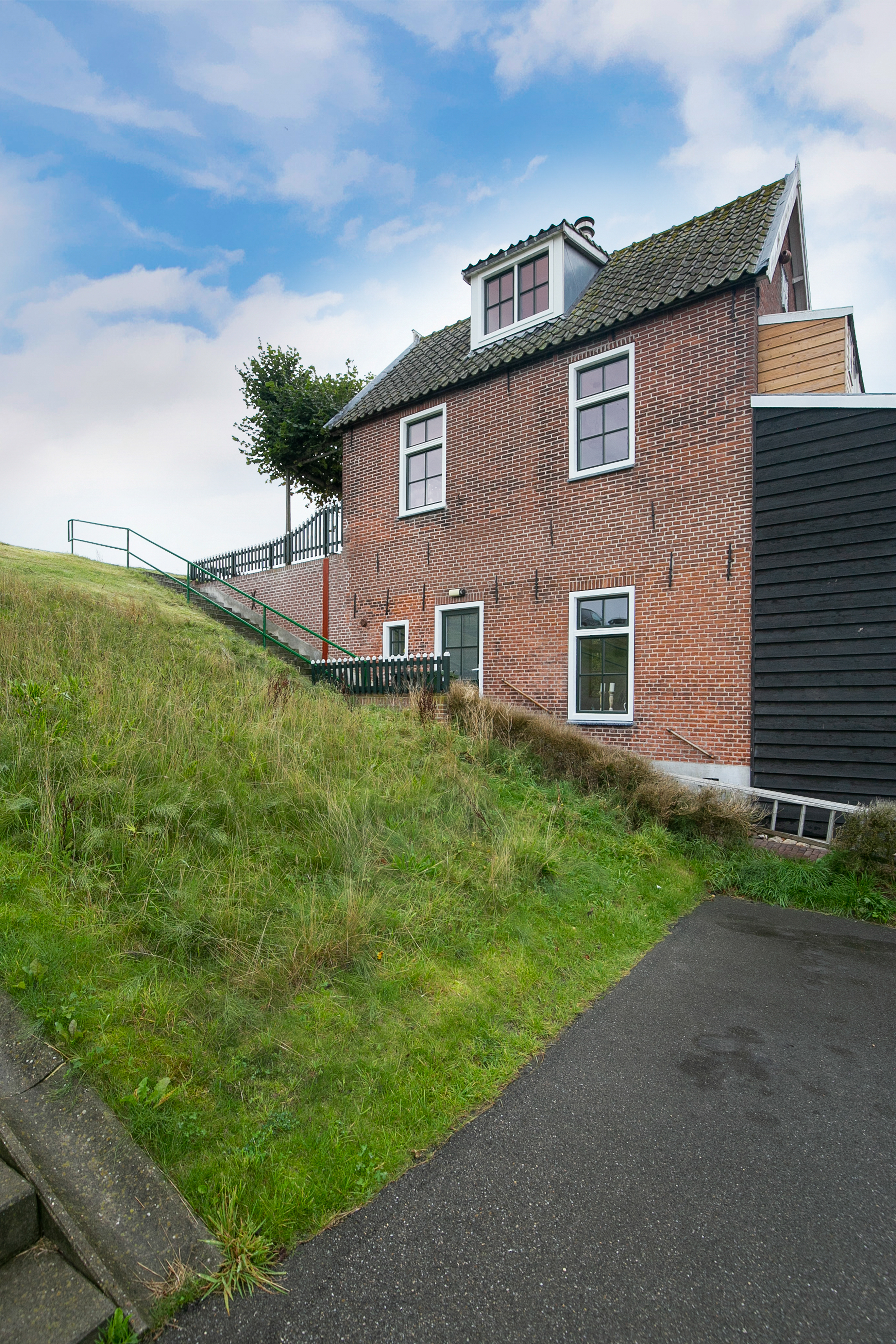 De afbeelding toont een traditioneel bakstenen huis met meerdere kozijnen en een deur. De kozijnen zijn symmetrisch geplaatst en hebben witte lijsten die afsteken tegen de rode baksteen. De deur bevindt zich op de begane grond en is ook in wit omlijst, wat goed past bij de kozijnen. Het huis heeft een klassieke, rustieke uitstraling, versterkt door het schuine dak en de omliggende groenvoorziening.