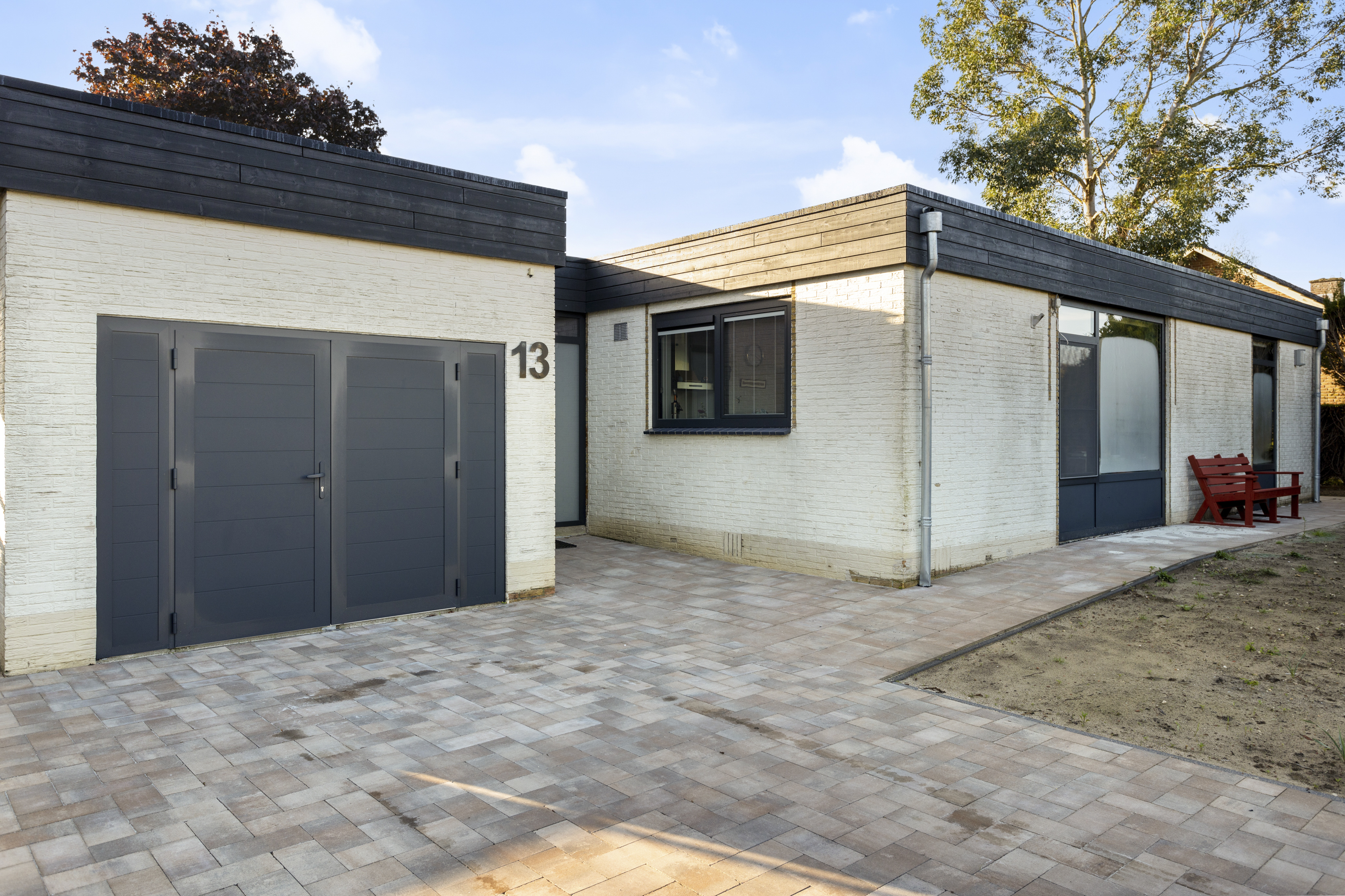De afbeelding toont een modern huis met een minimalistisch ontwerp. De gevel heeft grote, donker omlijste kozijnen en een bijpassende deur, wat een strak contrast biedt met de lichtgekleurde bakstenen muren. De kozijnen zijn rechthoekig en laten veel natuurlijk licht binnen. De deur is solide en past bij de algehele esthetiek van het huis.