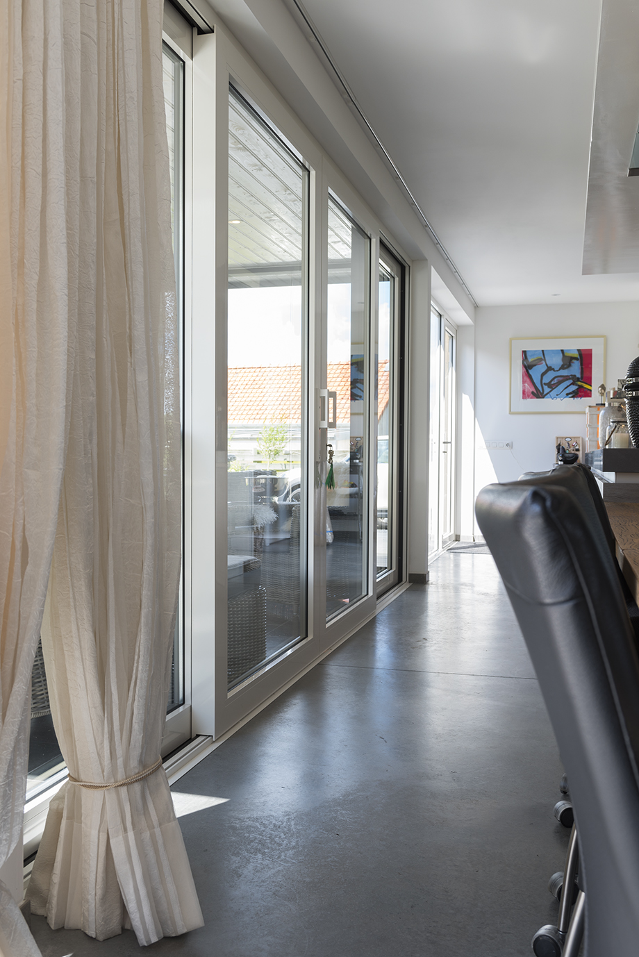 De afbeelding toont een modern interieur met grote schuifpuien die veel natuurlijk licht binnenlaten. De kozijnen zijn wit, wat goed past bij het minimalistische decor. Een set doorschijnende gordijnen voegt een vleugje elegantie toe, terwijl de gepolijste betonnen vloer de eigentijdse uitstraling versterkt.