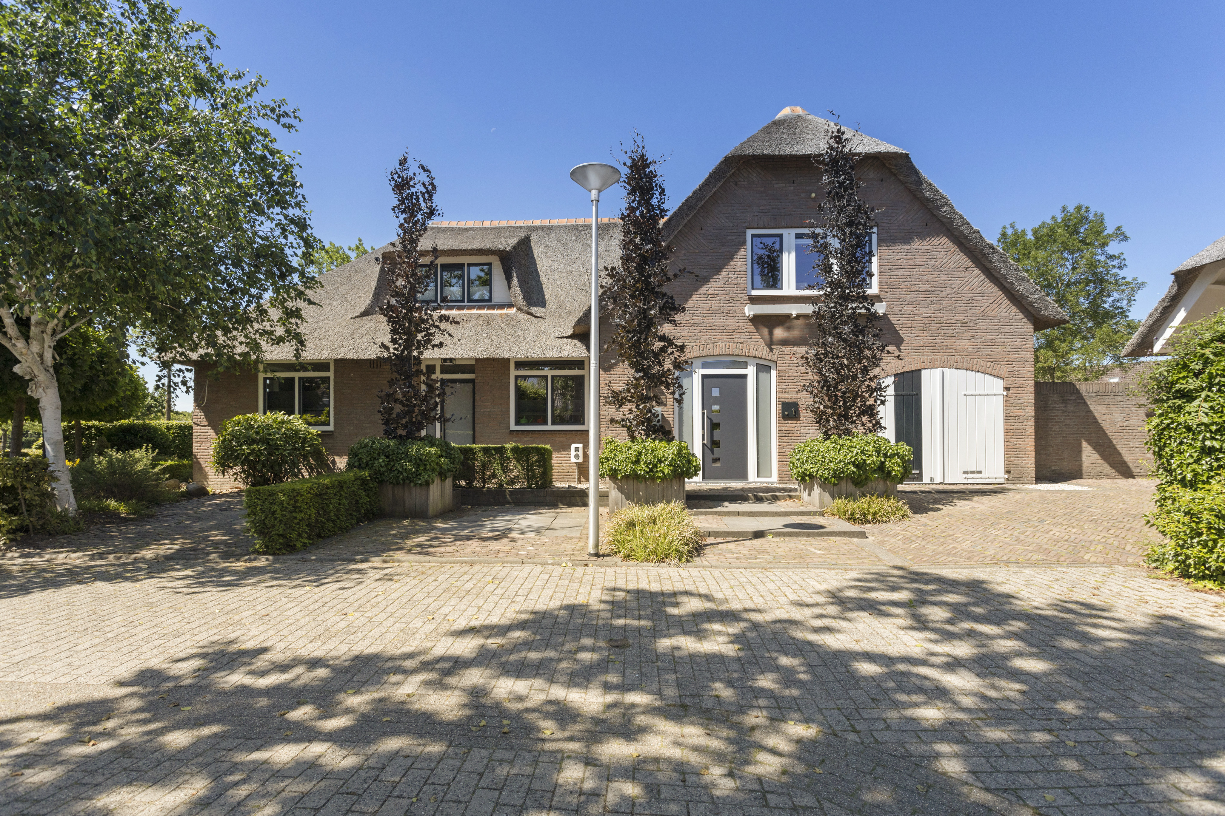 Het huis heeft een traditioneel rieten dak met moderne kozijnen en deuren. De kozijnen zijn rechthoekig met witte lijsten, wat contrasteert met de bakstenen gevel. De voordeur is strak en modern, met een metalen afwerking en verticale glazen panelen. De garagedeur is wit geschilderd, passend bij de kozijnen, en heeft een klassiek ontwerp.