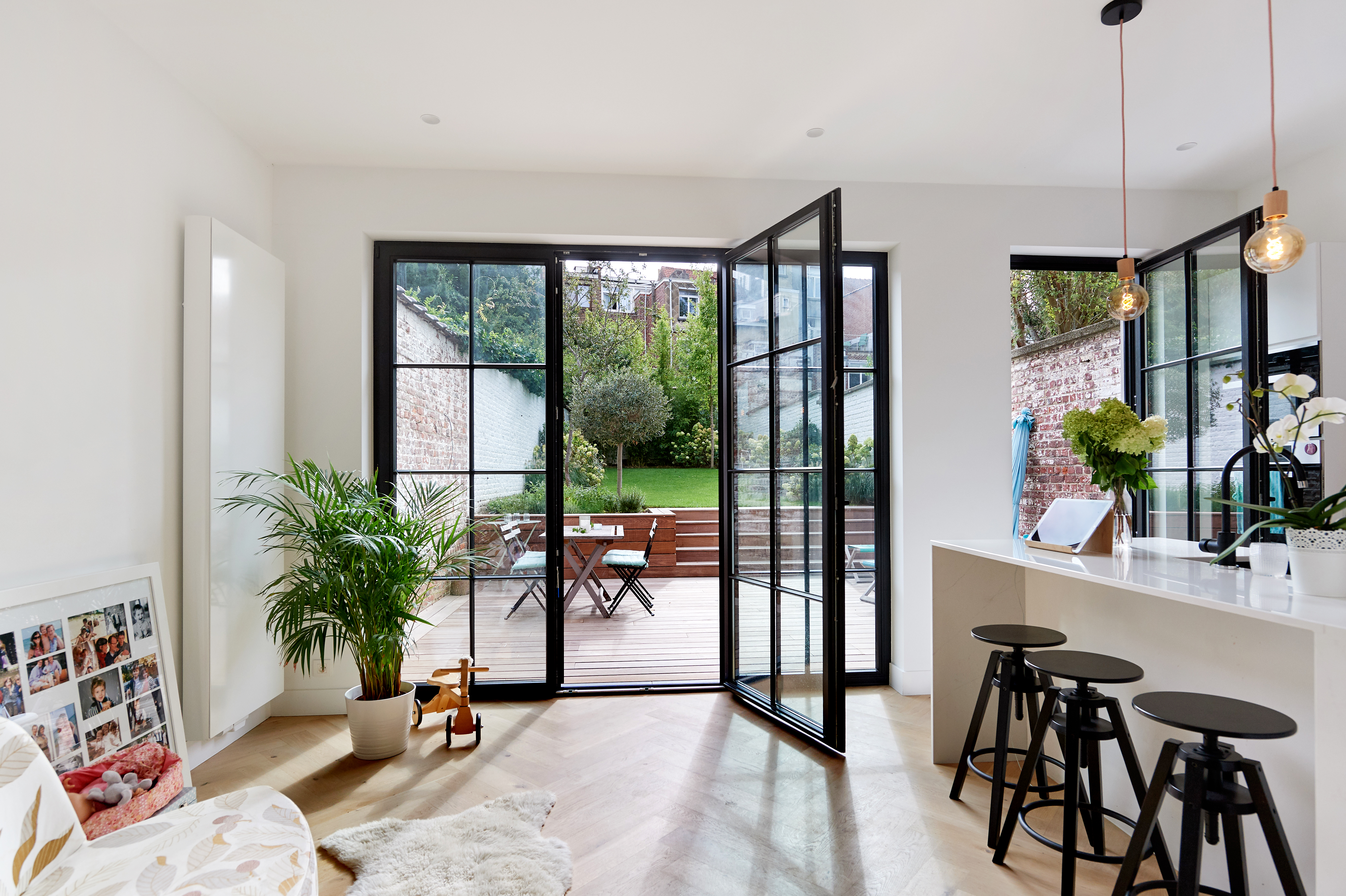 L'image montre un espace de vie moderne avec de grandes portes et châssis en verre encadrés de noir. Ces portes s'ouvrent sur une terrasse en bois et un jardin, laissant entrer la lumière naturelle. Le design épuré complète l'intérieur minimaliste, avec un îlot de cuisine et des tabourets de bar et des plantes décoratives.