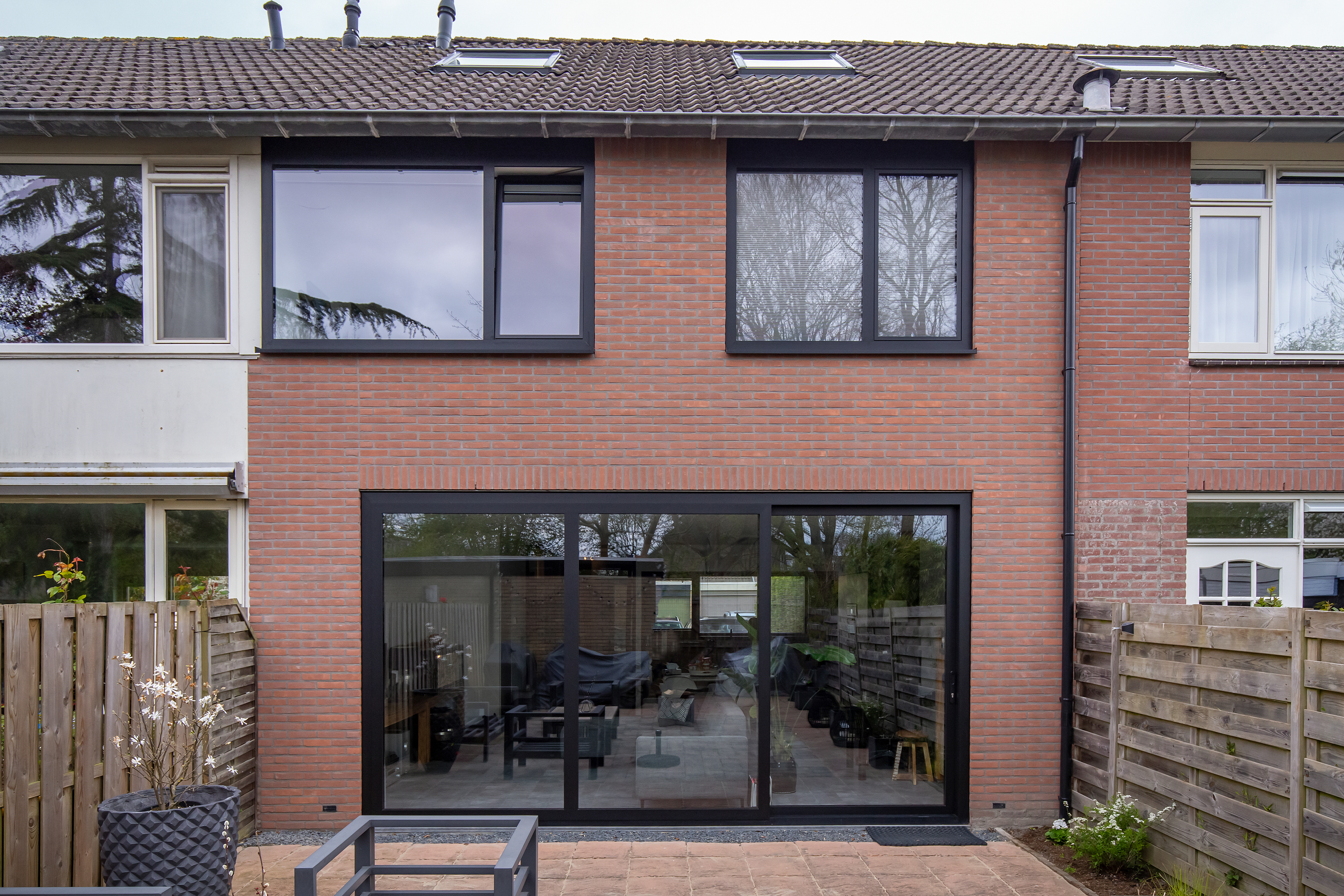 De afbeelding toont een modern bakstenen huis met grote ramen met zwarte kozijnen en een schuifpui. De bovenverdieping heeft twee brede ramen, terwijl de begane grond een grote schuifpui heeft die naar een terras leidt. Het ontwerp is strak en eigentijds, wat zorgt voor veel natuurlijk licht en uitzicht op buiten.