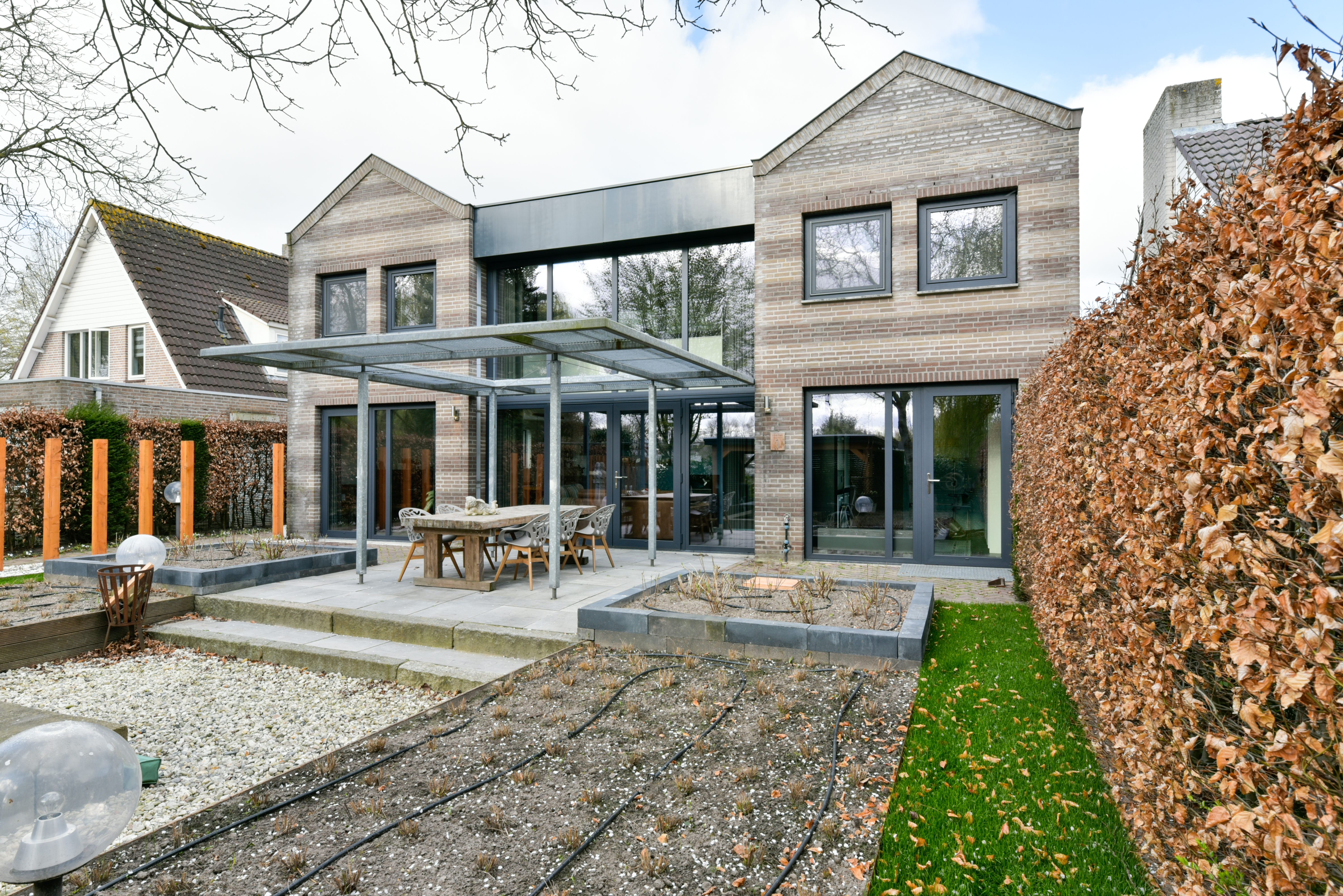 Het moderne huis heeft grote glazen schuifpuien en meerdere kozijnen, waardoor er veel natuurlijk licht binnenkomt. Het strakke ontwerp combineert baksteen en glas, met de deuren die openen naar een ruime patio.