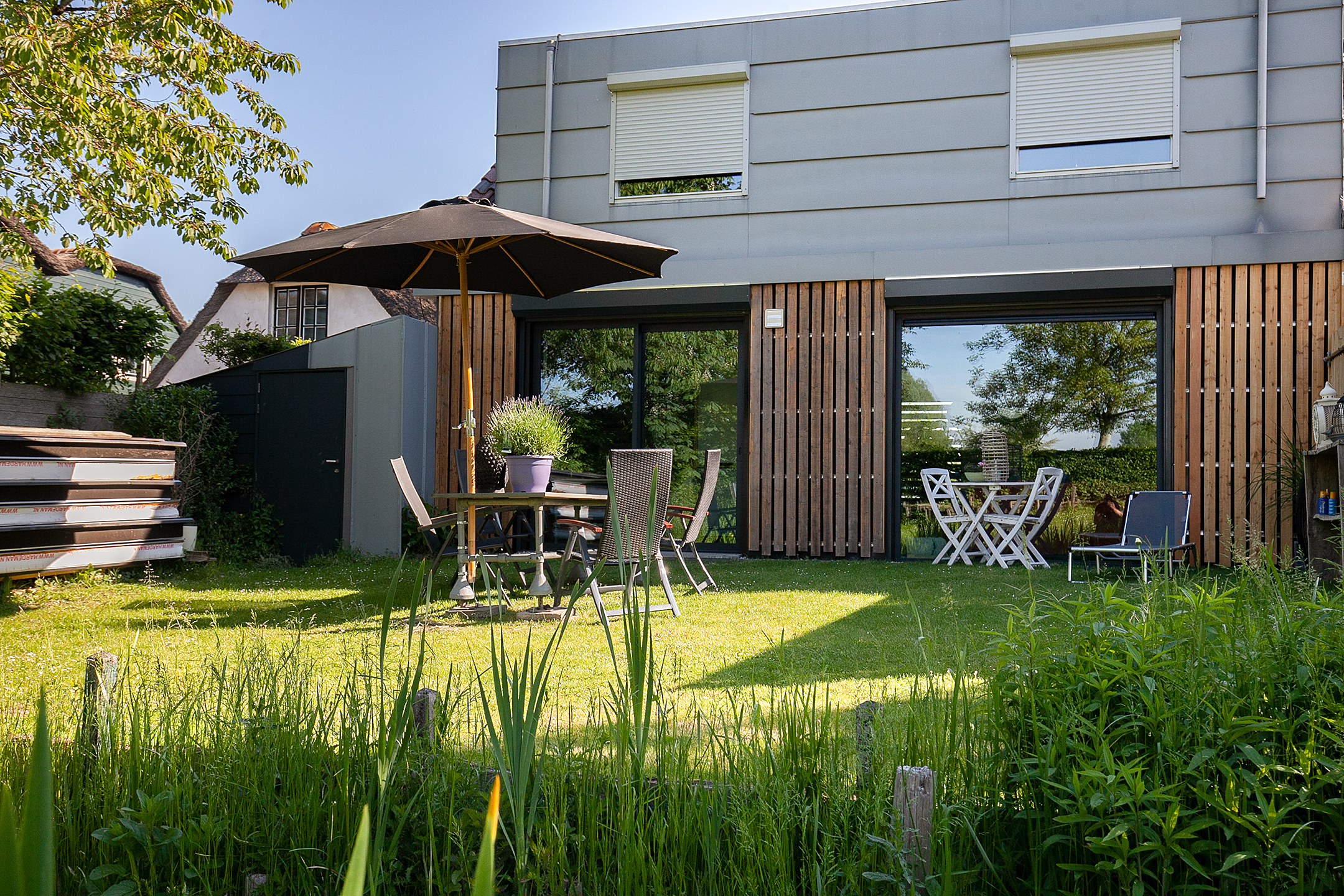 Het moderne huis heeft grote schuifpuien die uitkijken op een weelderige tuin. De kozijnen zijn voorzien van strakke zwarte randen, die mooi passen bij de houten panelen aan de buitenkant. Boven zijn er kleinere kozijnen met rolluiken voor extra licht en ventilatie. De deuren zijn geïntegreerd in de schuifpuien, wat een naadloze toegang tot de buitenruimte biedt.