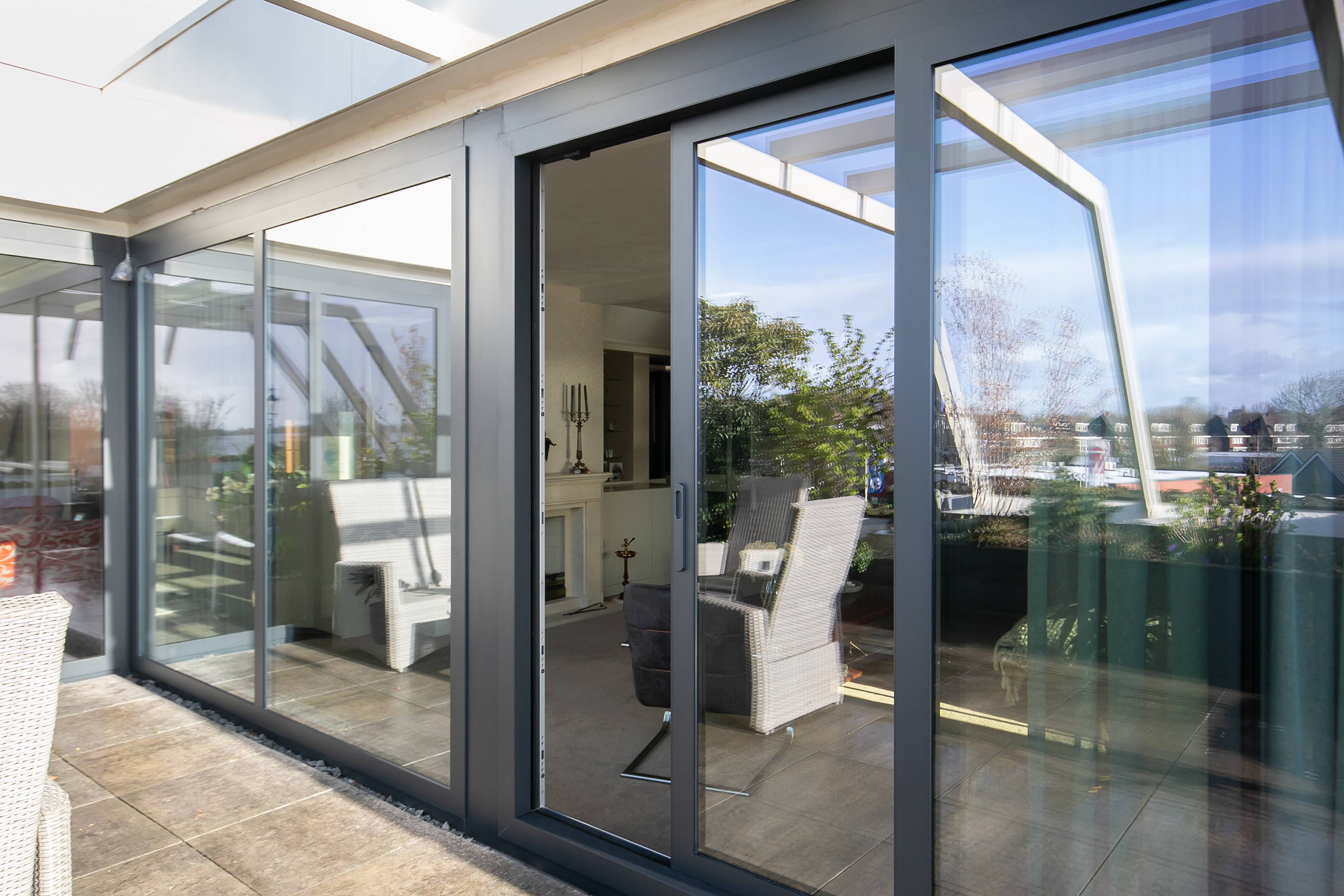 De afbeelding toont een moderne set schuifpuien die naar een terras leiden. De puien zijn omlijst met strak, donker metaal, wat een eigentijdse uitstraling geeft. De grote glasplaten laten veel natuurlijk licht binnen, waardoor er een naadloze verbinding tussen binnen en buiten ontstaat.