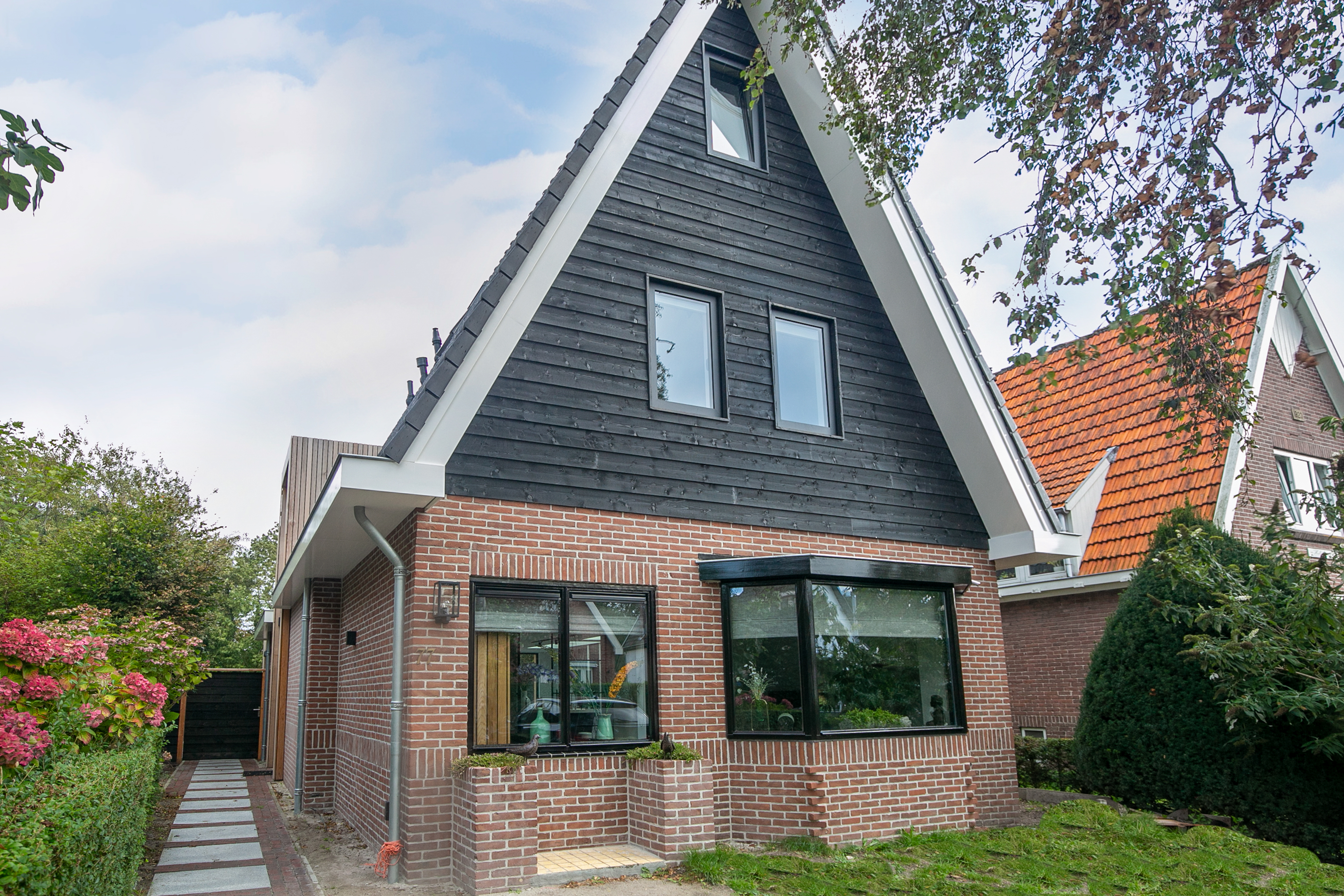 Het huis heeft een modern ontwerp met een combinatie van baksteen en donker hout. De voorgevel bevat grote kozijnen die veel natuurlijk licht binnenlaten. De kozijnen zijn in het zwart omlijst, wat bijdraagt aan de strakke uitstraling. De voordeur is subtiel geïntegreerd in het ontwerp, waardoor de strakke lijnen van de architectuur behouden blijven.