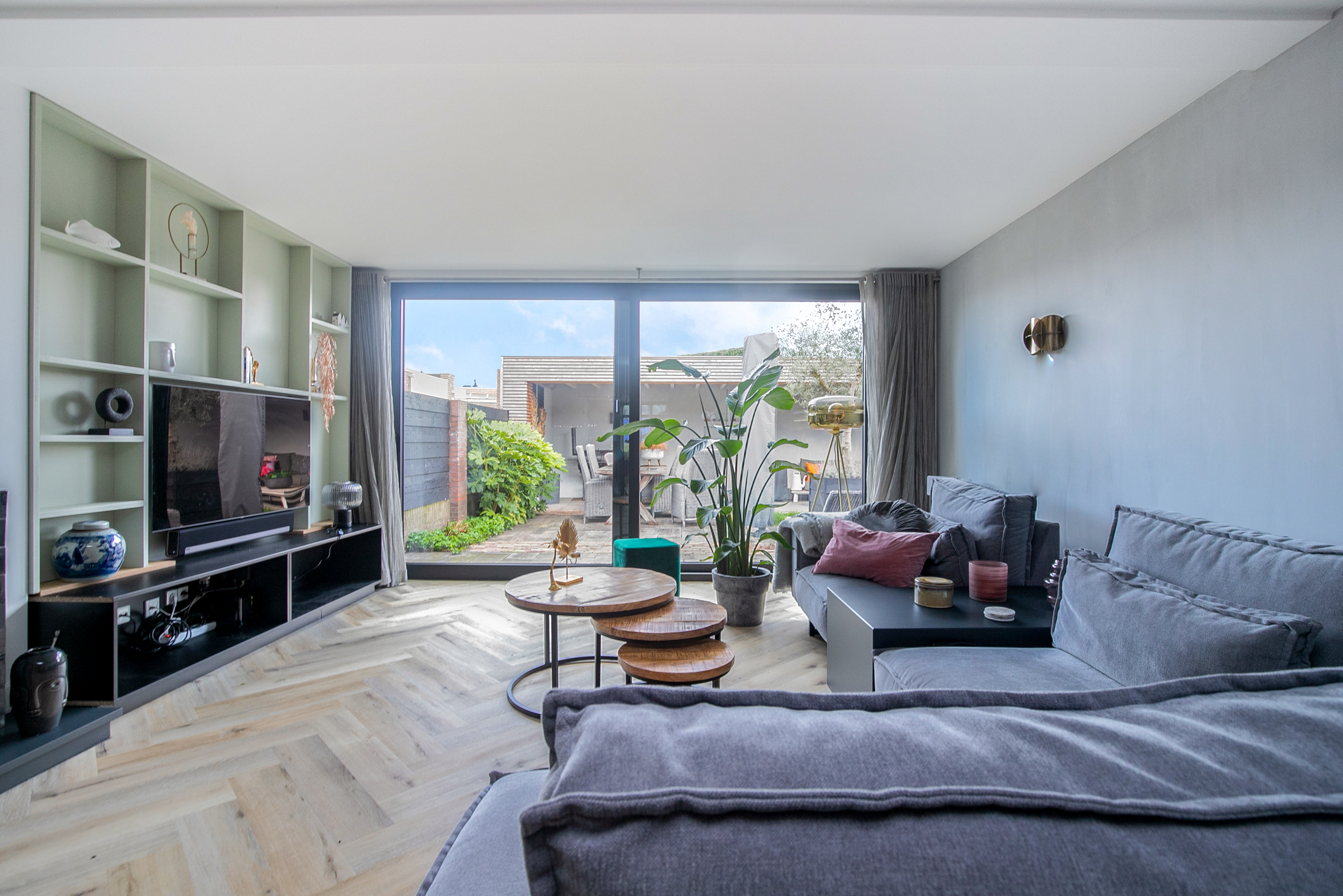De afbeelding toont een moderne woonkamer met grote schuifpuien die uitkijken op een tuin. De kozijnen laten veel natuurlijk licht binnen, wat zorgt voor een heldere en luchtige sfeer. Het strakke ontwerp van de kozijnen past perfect bij het eigentijdse interieur van de kamer.