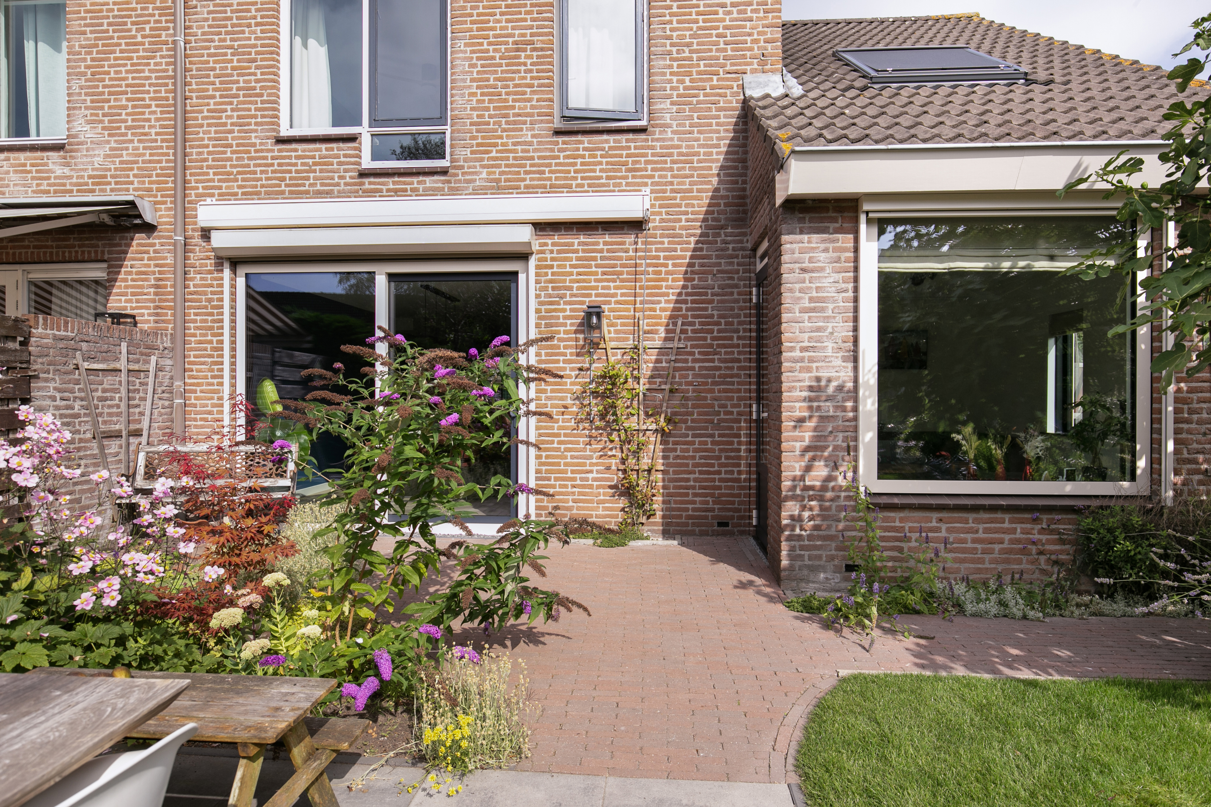 De afbeelding toont een bakstenen huis met grote kozijnen en een schuifpui. De kozijnen zijn modern en laten veel licht binnen. De schuifpui opent naar een tuin met kleurrijke bloemen.