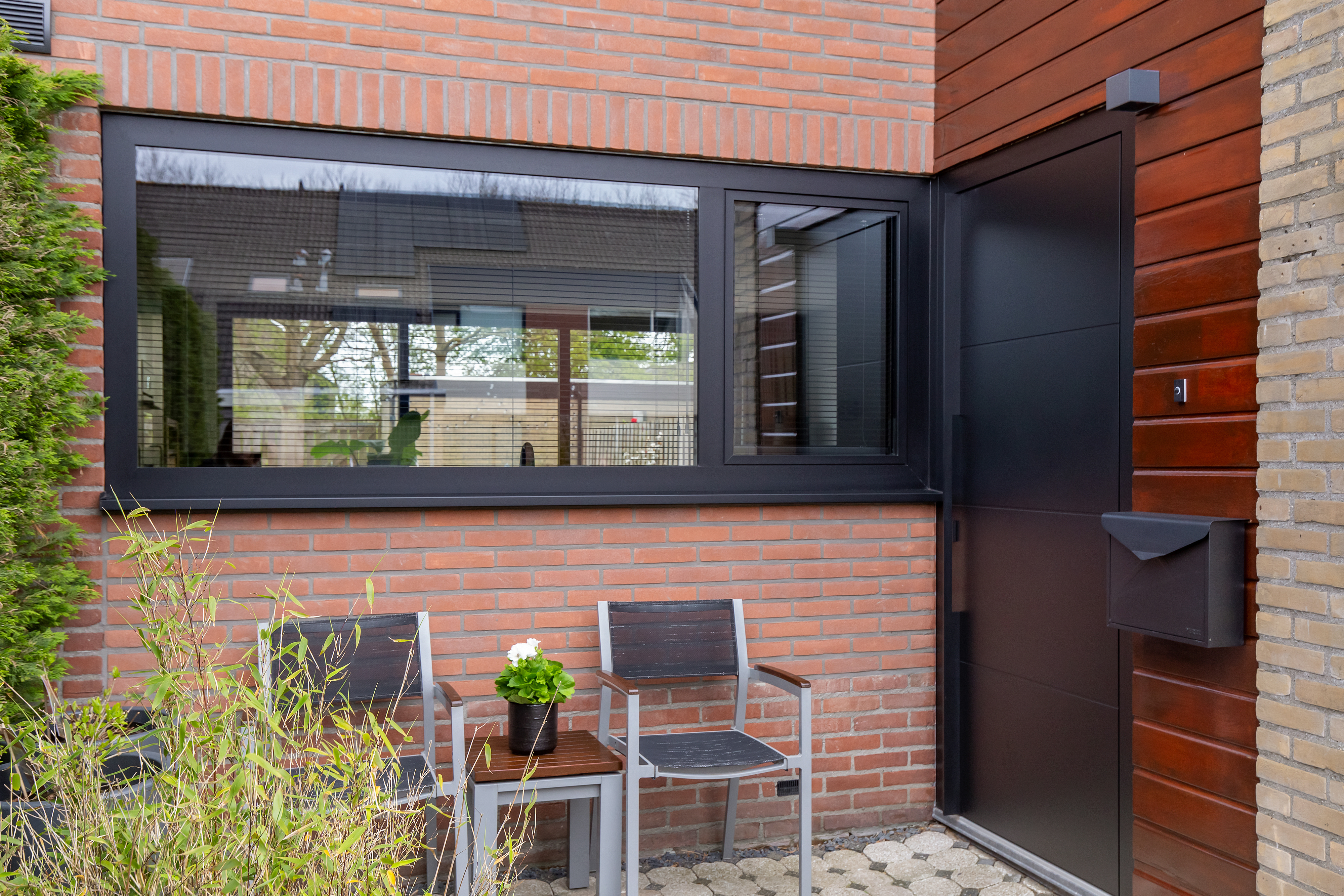 De afbeelding toont een moderne huisgevel met een groot zwart omlijst kozijn en een strakke zwarte deur. Het kozijn weerspiegelt de omgeving, wat diepte toevoegt aan de scène. De deur is stevig en past bij de bakstenen en houten gevel.