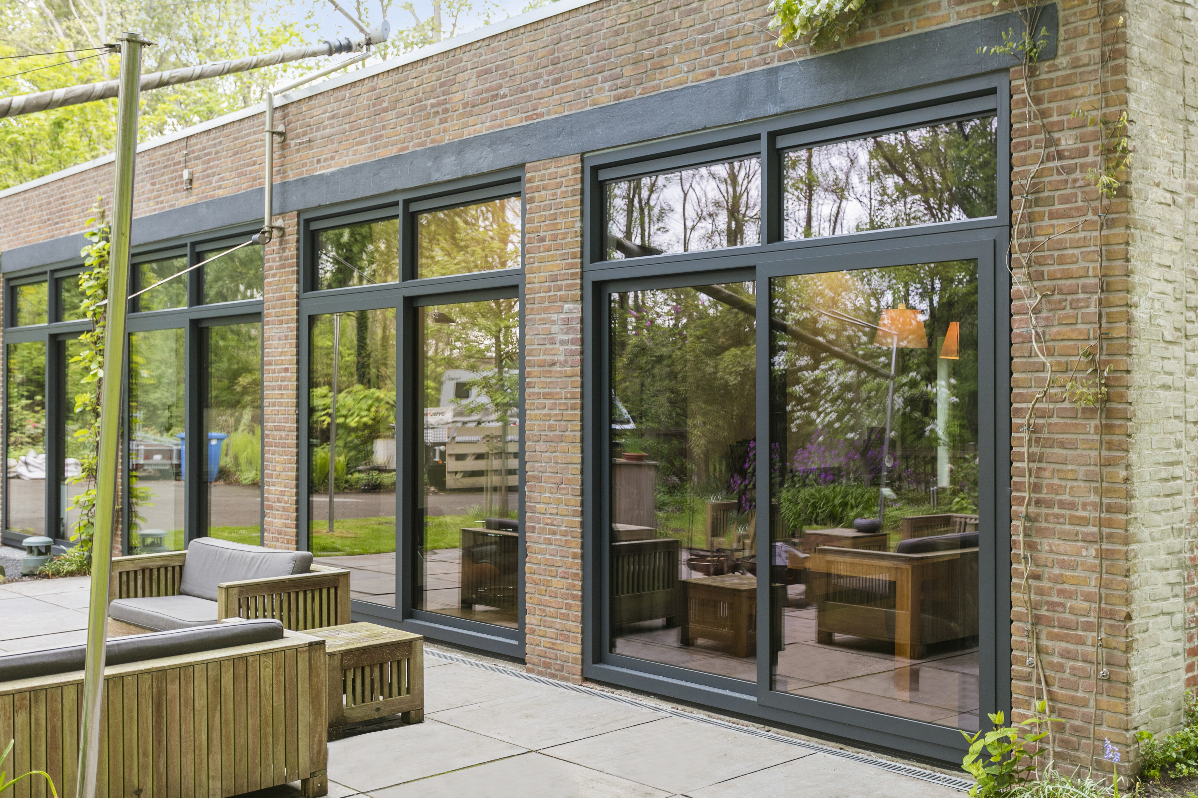 De afbeelding toont een moderne gevel met grote glazen schuifpuien en kozijnen. De zwarte kozijnen vormen een mooi contrast met de bakstenen buitenkant. Het glas weerspiegelt de omliggende natuur, wat zorgt voor een naadloze overgang tussen binnen en buiten.