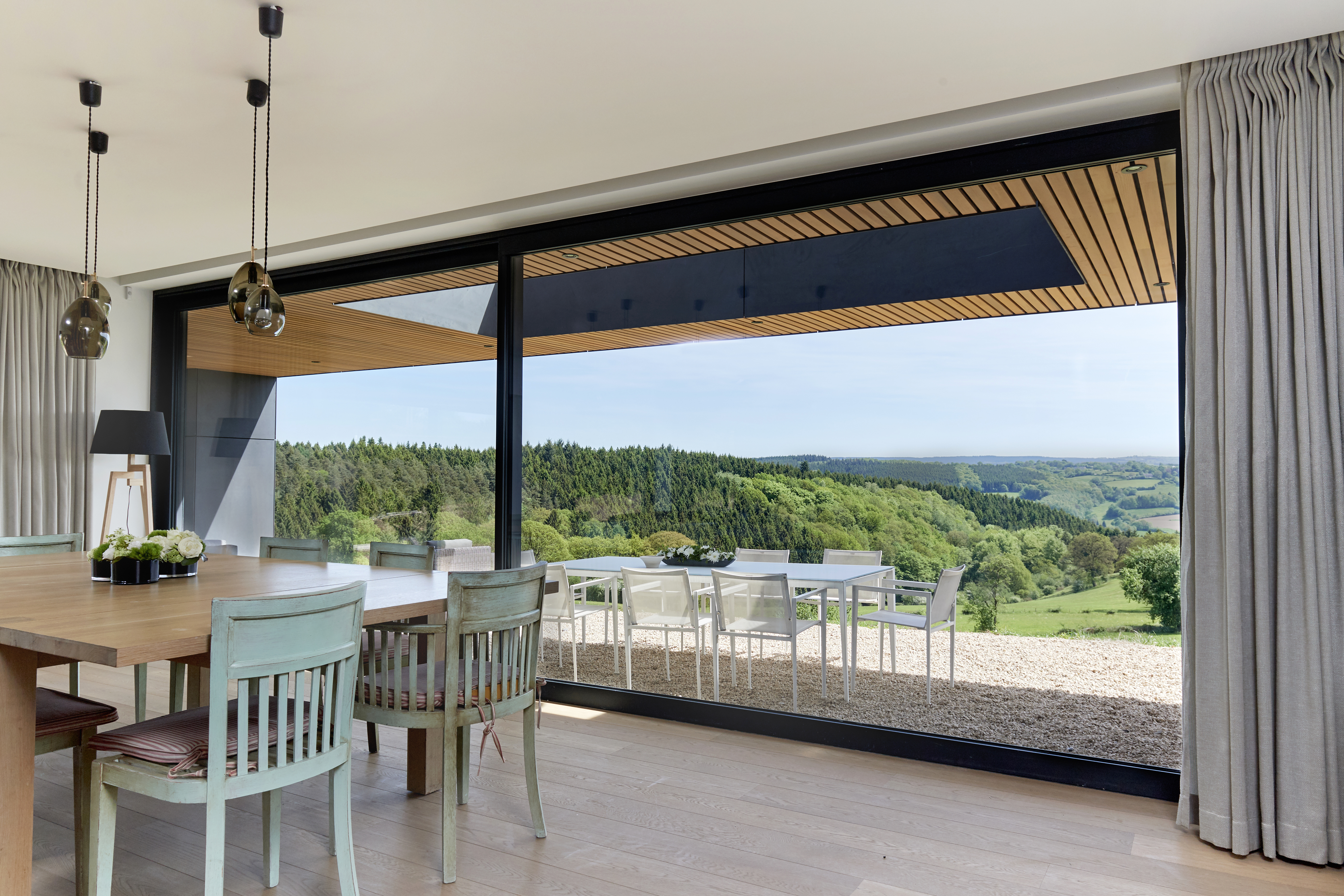 L'image montre un intérieur moderne avec de grands châssis coulissants offrant une vue panoramique sur un paysage verdoyant. Les châssis sont encadrés de noir élégant, complétant le décor minimaliste. Les portes coulissantes s'ouvrent sur une terrasse avec des sièges extérieurs, fusionnant harmonieusement les espaces intérieurs et extérieurs.