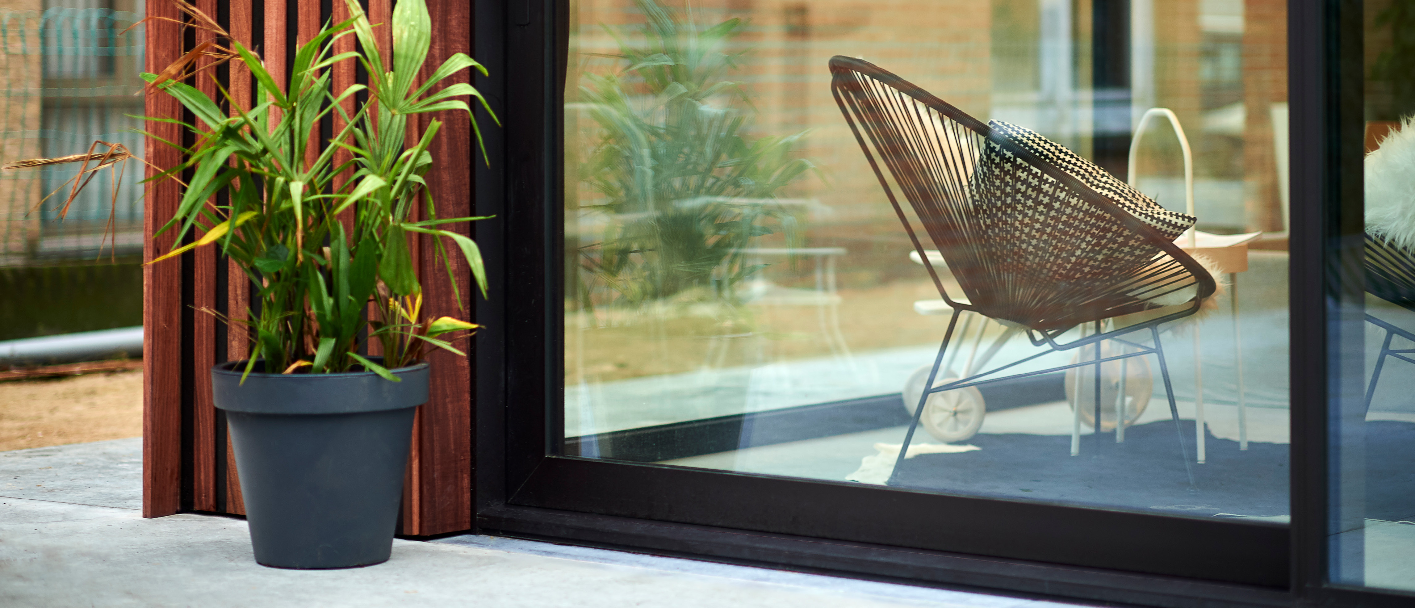 De afbeelding toont een modern schuifraam dat een naadloze overgang biedt tussen binnen- en buitenruimtes. Het strakke zwarte frame van het raam past bij het eigentijdse ontwerp en laat natuurlijk licht binnenstromen. Een potplant en een stijlvolle stoel zijn zichtbaar door het glas, wat de esthetische aantrekkingskracht vergroot.