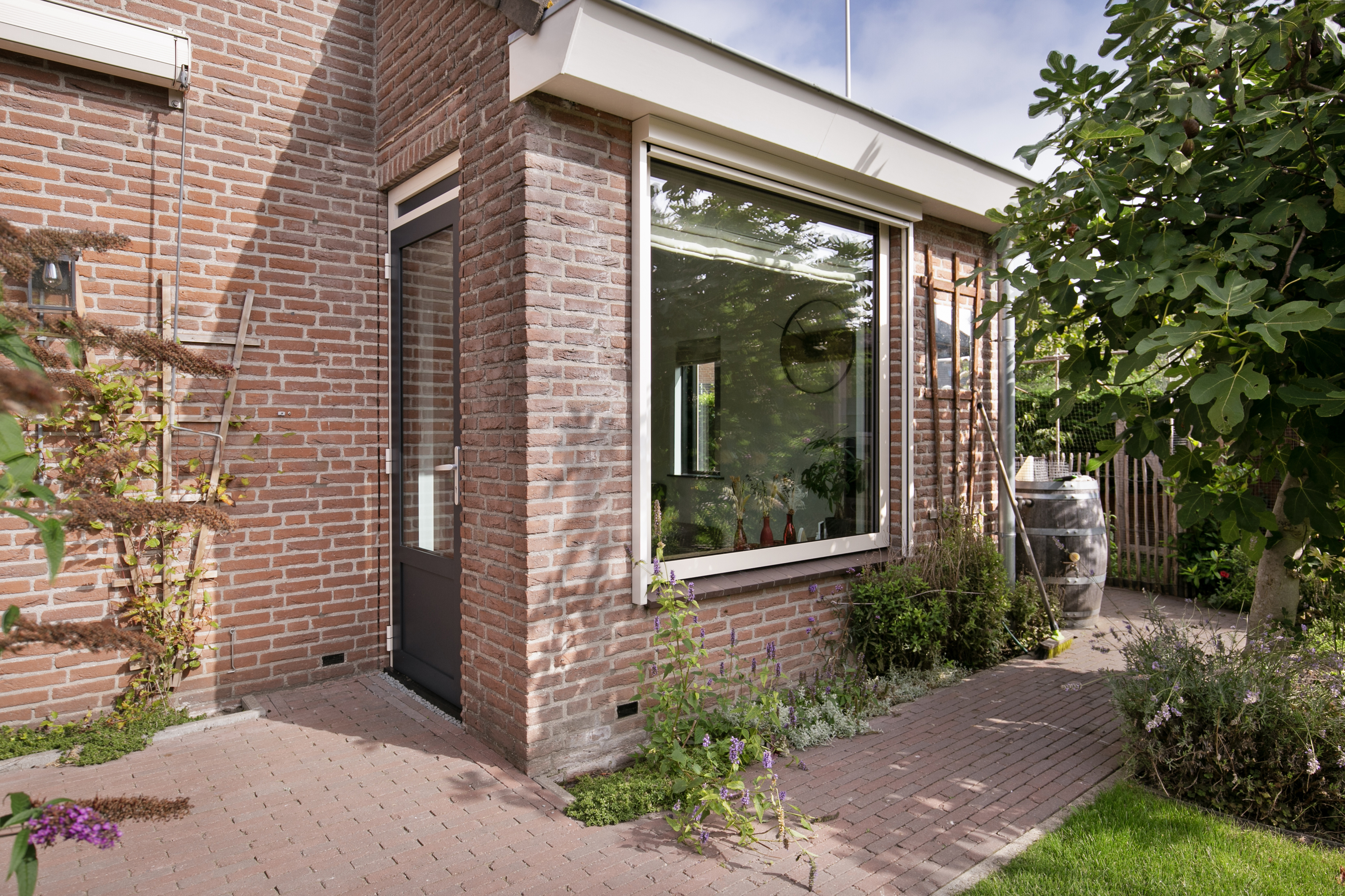 De afbeelding toont een hoek van een bakstenen huis met een groot kozijn en een glazen deur. Het kozijn is wit omlijst en laat natuurlijk licht binnen. De deur is ook wit omlijst, wat mooi bij het kozijn past. Het metselwerk geeft een rustieke charme en de omliggende tuin versterkt de gezellige sfeer.