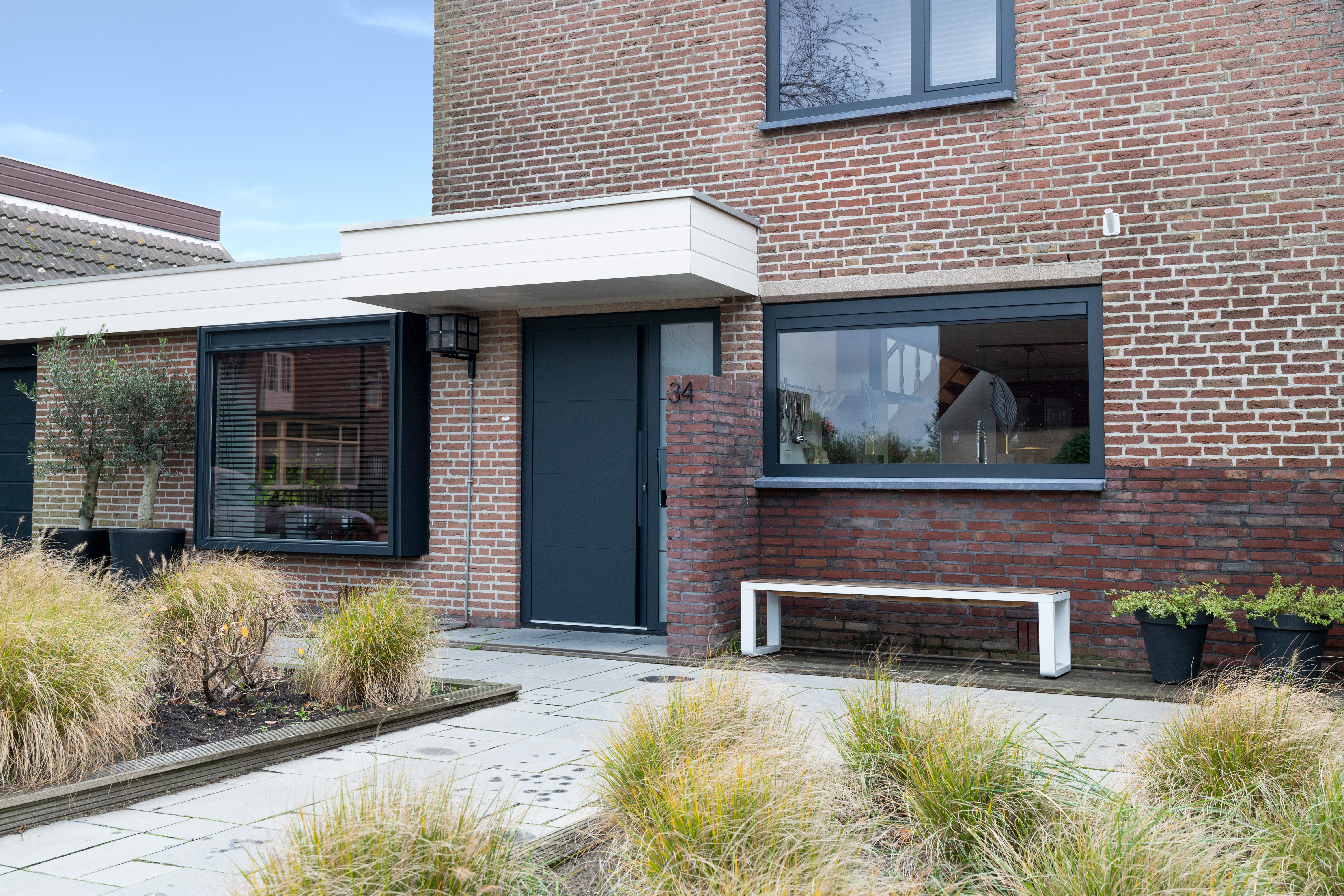 De afbeelding toont een modern bakstenen huis met grote, donker omlijste kozijnen en een strakke, bijpassende deur. De kozijnen zijn breed en laten veel licht binnen. De deur is centraal geplaatst en past bij het eigentijdse ontwerp van het huis.