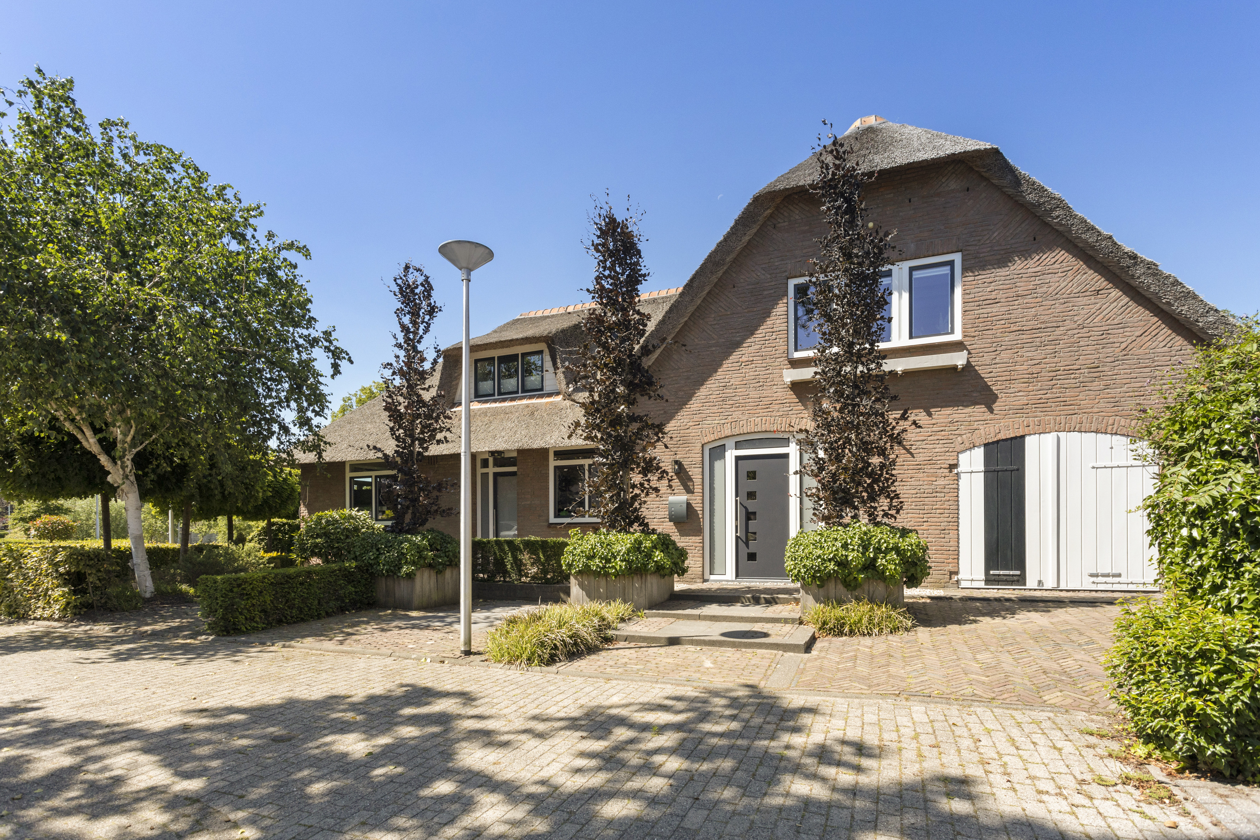 Het huis heeft een traditionele uitstraling met een rieten dak en een bakstenen gevel. De kozijnen zijn modern, met witte lijsten die afsteken tegen de baksteen. De voordeur is strak en eigentijds, met een reeks kleine vierkante raampjes. Luiken op de onderste kozijnen geven een rustieke toets.