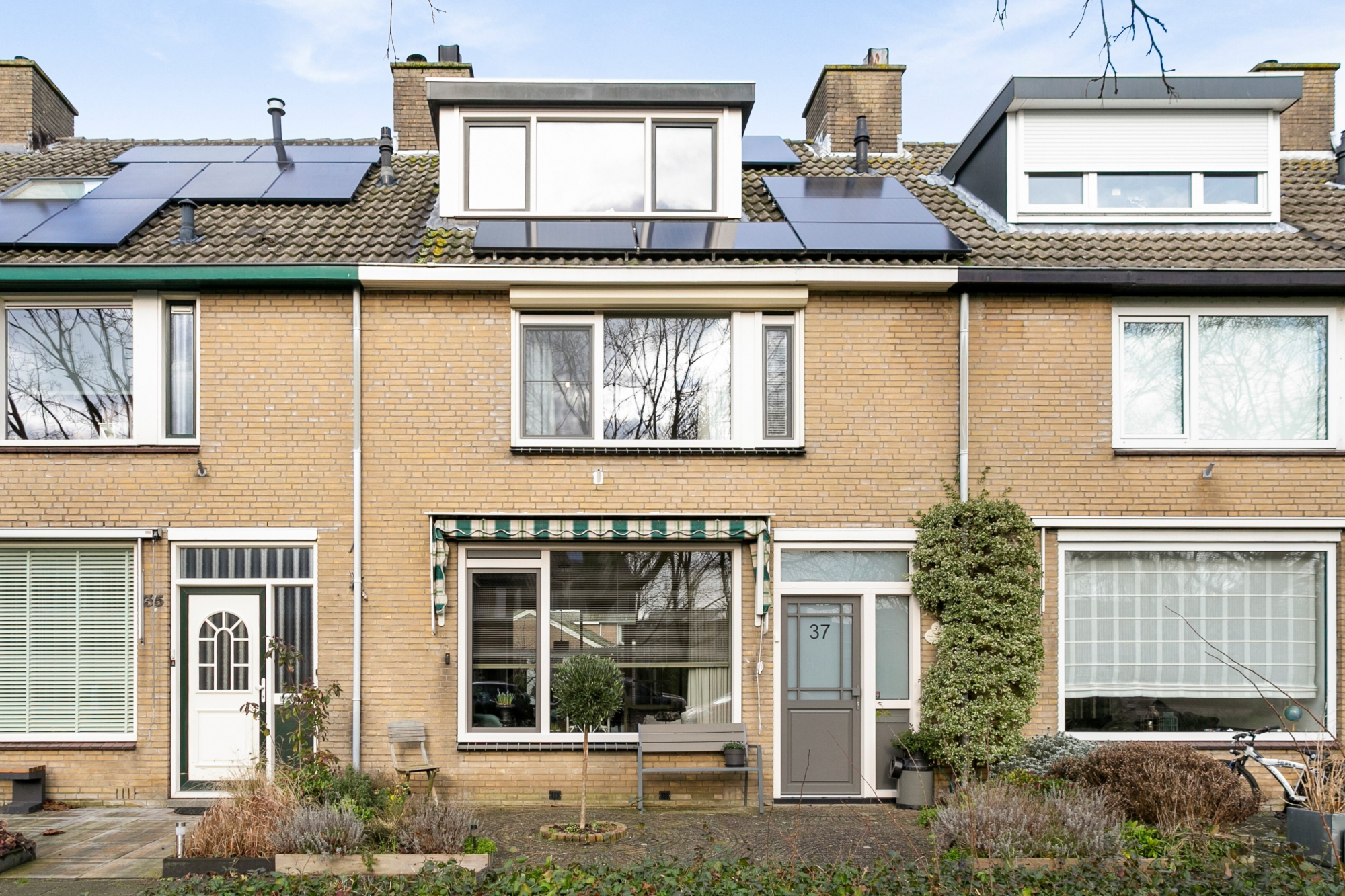 De afbeelding toont een rij bakstenen huizen met grote kozijnen en deuren. Elk huis heeft een combinatie van moderne en traditionele elementen, met grote glazen kozijnen die natuurlijk licht binnenlaten. De deuren zijn eenvoudig maar elegant, passend bij de algehele esthetiek van de woningen.