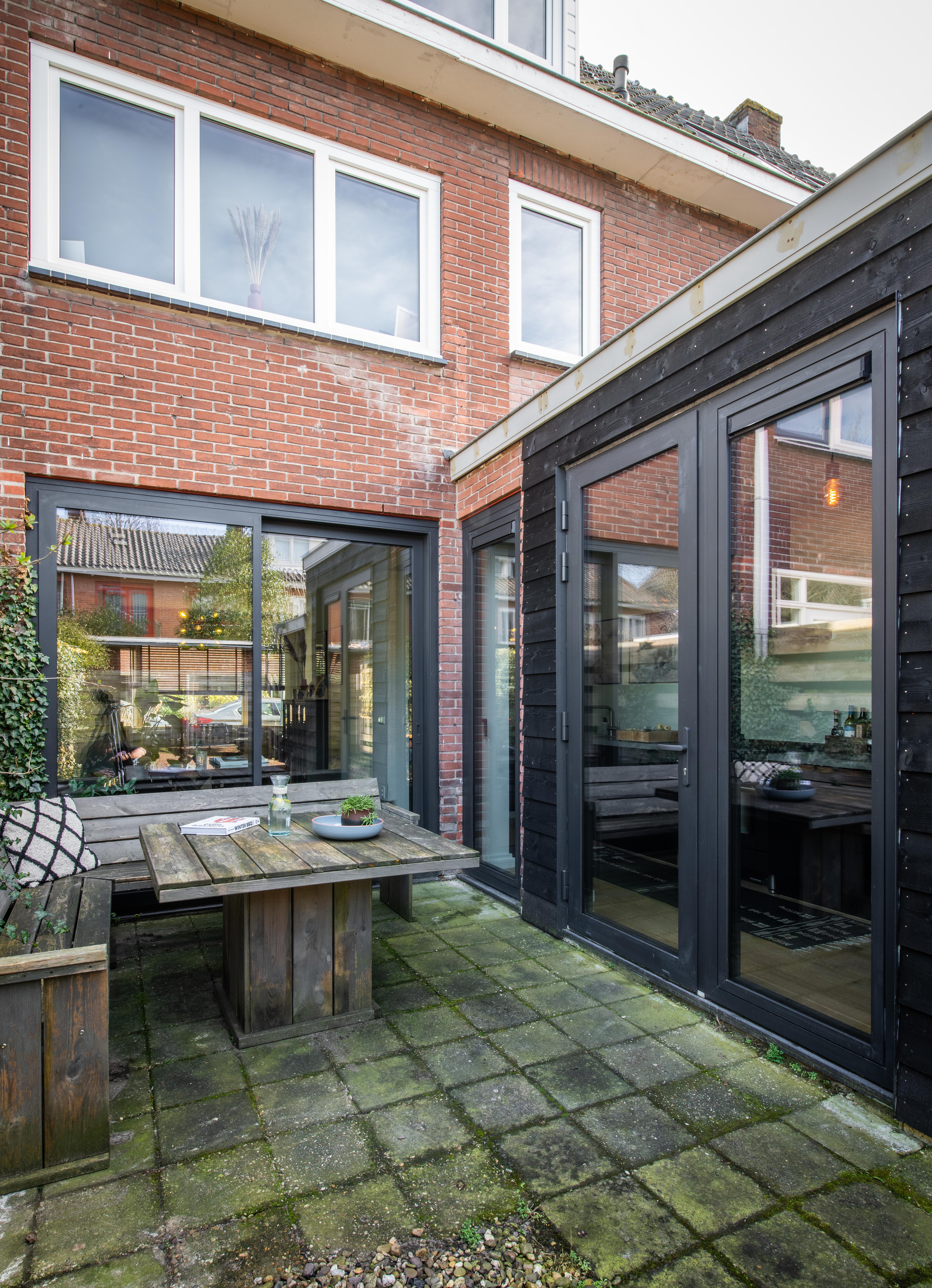 De afbeelding toont een moderne huisgevel met grote glazen schuifpuien en meerdere kozijnen. De schuifpuien hebben een donker metalen frame, wat een strak contrast biedt met de bakstenen muren. De kozijnen boven zijn rechthoekig en symmetrisch uitgelijnd, wat het architectonische ontwerp versterkt.