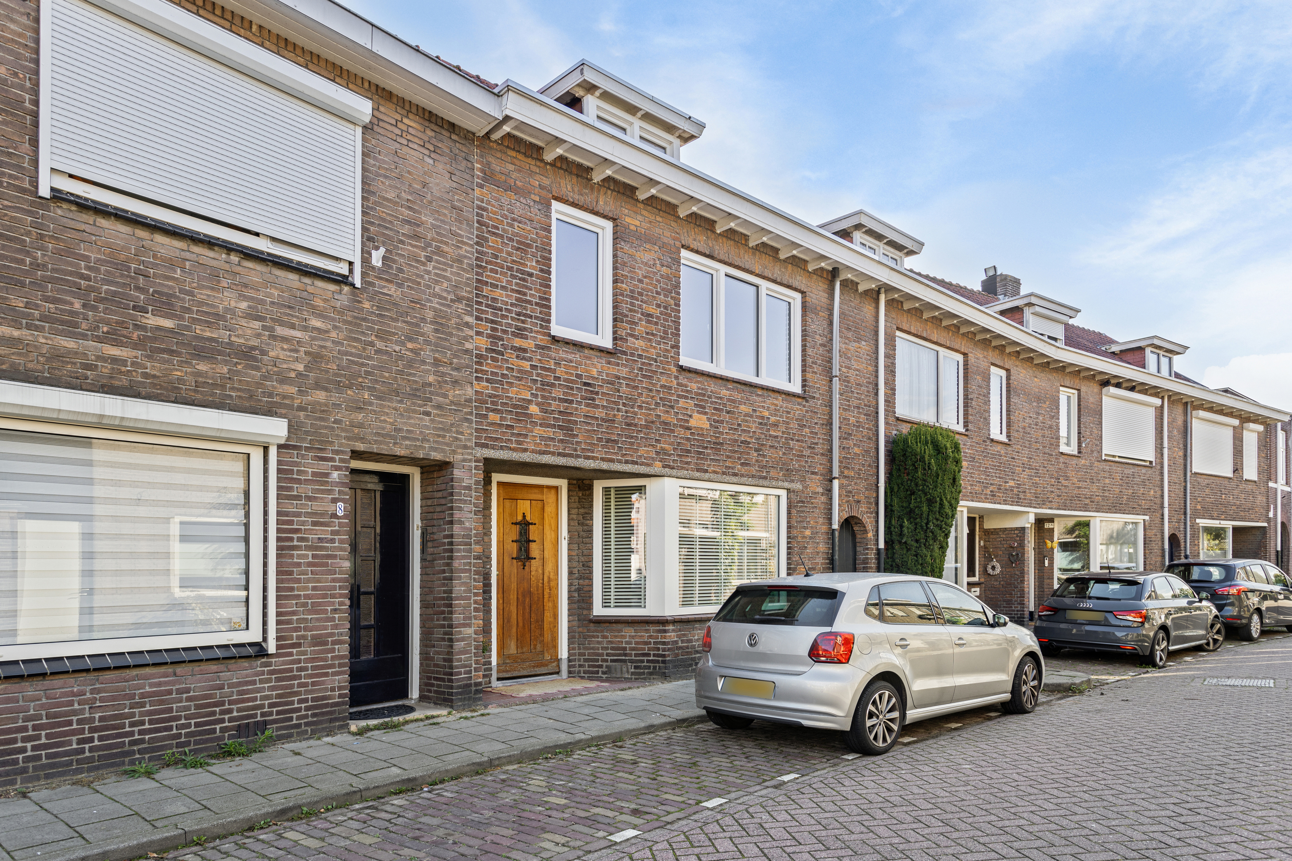 De afbeelding toont een rij bakstenen huizen met grote rechthoekige kozijnen en houten deuren. De kozijnen hebben witte lijsten en sommige hebben rolluiken. De deuren zijn van massief hout met decoratieve elementen. De straat is vol geparkeerde auto's.