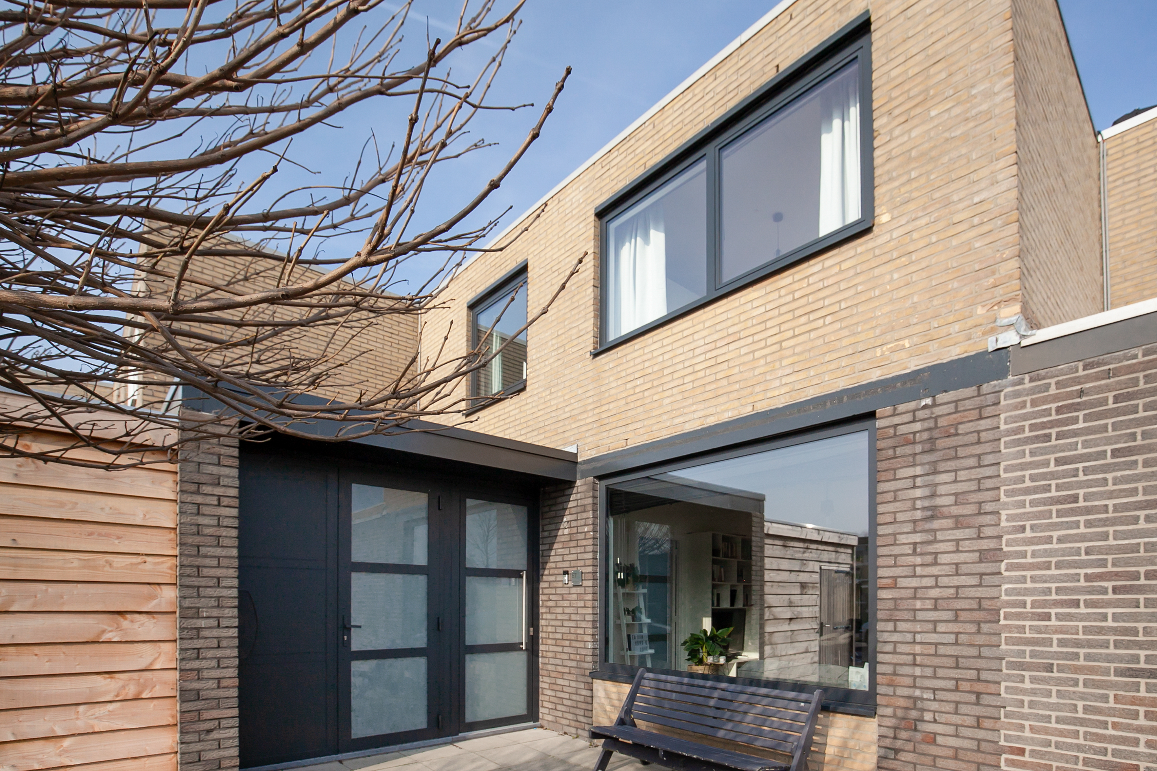 Het moderne huis heeft grote, zwartomrande kozijnen en een strakke zwarte deur. De kozijnen zijn prominent aanwezig op zowel de begane grond als de bovenverdieping, waardoor er veel natuurlijk licht binnenkomt. De deur is dubbel, met matglazen panelen, wat het eigentijdse ontwerp complimenteert.