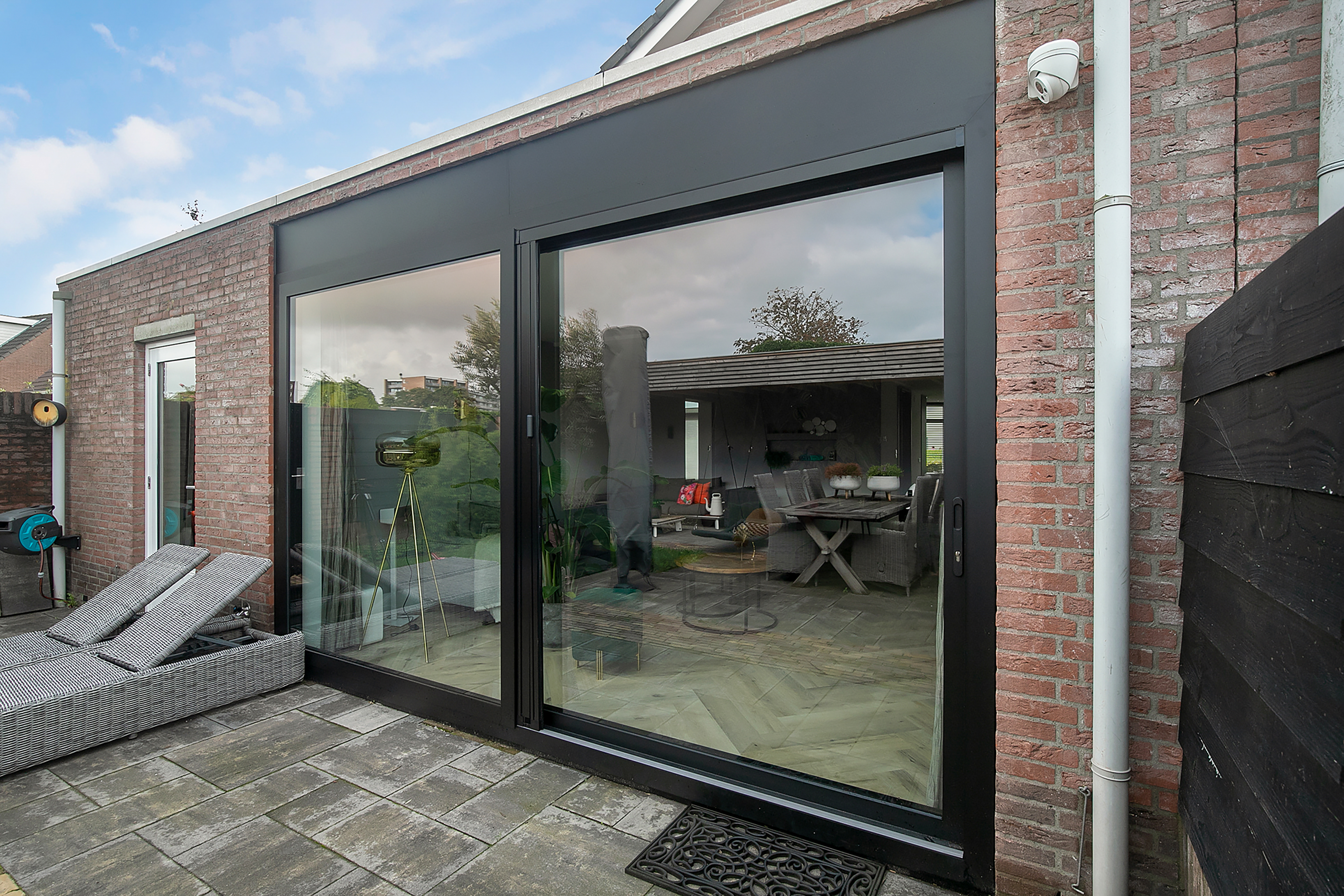 De afbeelding toont een moderne woning met grote schuifpuien en een kleinere zijdeur. De schuifpuien zorgen voor een naadloze overgang tussen binnen en buiten, waardoor natuurlijk licht de binnenruimte overspoelt. De puien hebben een strak zwart frame dat mooi contrasteert met de bakstenen gevel. Het terras heeft comfortabele zitplaatsen, wat de buitenervaring verrijkt.