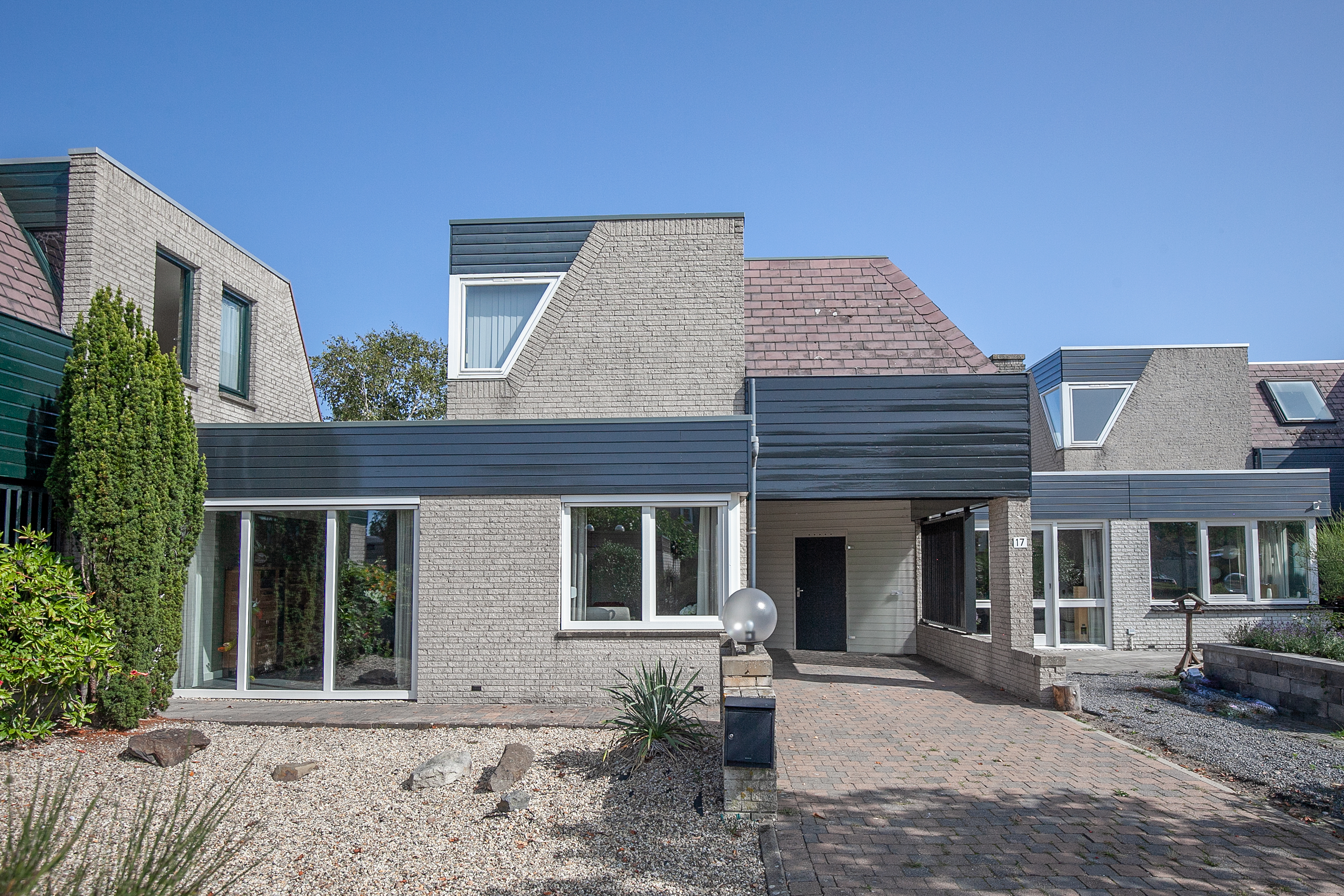 Het moderne huis heeft grote schuifpuien en een combinatie van rechthoekige en driehoekige kozijnen, wat veel natuurlijk licht binnenlaat. De voordeur is eenvoudig en past bij het strakke ontwerp van de gevel.