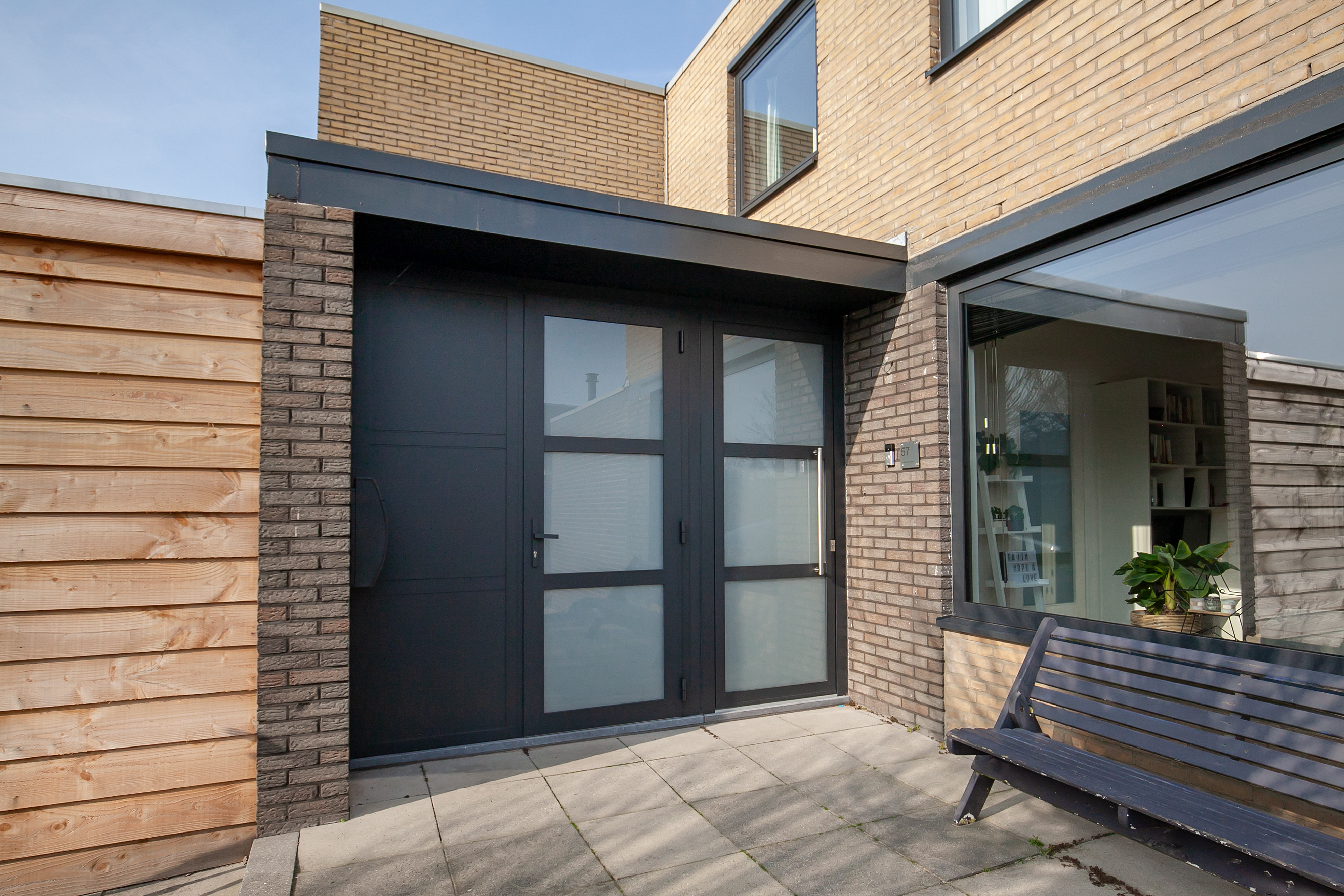 De afbeelding toont een moderne huisingang met grote glazen deuren en kozijnen. De deuren zijn omlijst met donker metaal, wat een strakke en eigentijdse uitstraling geeft. Naast de deuren zijn er grote kozijnen die natuurlijk licht binnenlaten. De combinatie van baksteen en hout op de buitenmuren zorgt voor een warme en uitnodigende sfeer.