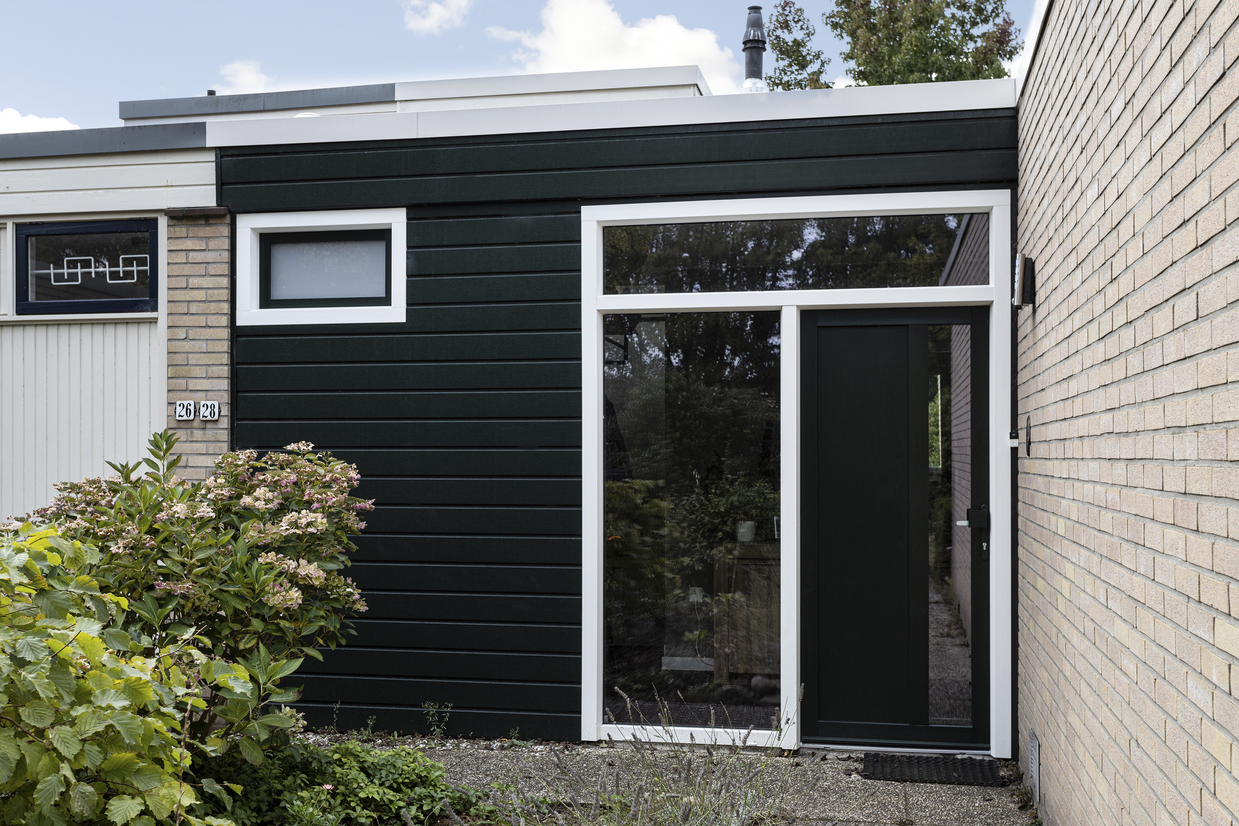 De afbeelding toont een moderne gevel van een huis met een donkergroene deur en een bijpassend kozijn. De deur is strak en eigentijds, met een groot glazen paneel dat natuurlijk licht binnenlaat. Het kozijn is rechthoekig en boven de deur geplaatst, wat extra licht en een samenhangend ontwerp biedt. De gevel wordt aangevuld door lichte bakstenen en een kleine tuin.